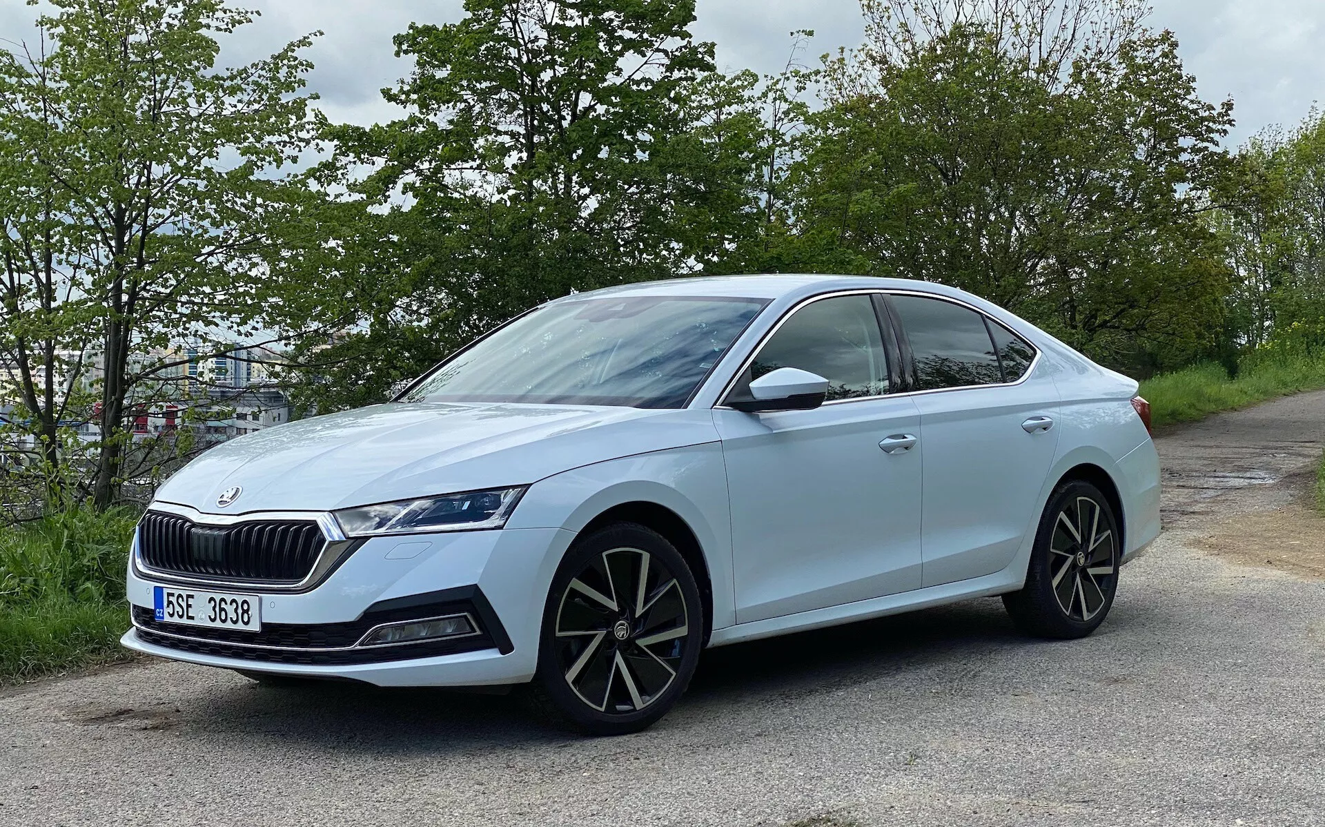 Umiarkowanie kątowy widok boczny jasnoniebieskiego sedana Skoda Octavia 2020, umieszczonego przy drodze z zielonym listowiem w tle. Prezentuje nowoczesny design z elegancką, czterodrzwiową nadwoziem, wyraźnym grillem, agresywnymi reflektorami LED, dużymi, wieloszprychowymi felgami aluminiowymi, lusterkami bocznymi w kolorze nadwozia z wbudowanymi kierunkowskazami, opadającą linią dachu, przyciemnionymi szybami z chromowanymi listwami, drzwi z wbudowanymi klamkami, oponami niskoprofilowymi i oznaczeniem wskazującym model lub pakiet technologiczny.