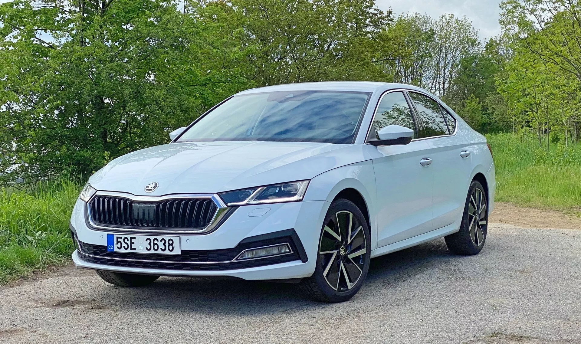 Obraz jasno-niebiesko-szarej Škody Octavia 2020 ustawionej na asfaltowej drodze z zielenią w tle. Zdjęcie wykonane pod kątem trzech czwartych od przodu podkreśla detale designu: wyraźny przedni grill z pionowymi listwami i emblematem Škody, eleganckie, kątowe reflektory LED z światłami do jazdy dziennej, oddzielne światła przeciwmgielne, duże, wieloszprychowe felgi aluminiowe, lusterka boczne z wbudowanymi kierunkowskazami, prawdopodobnie przednie czujniki parkowania oraz dynamiczne linie nadwozia wzdłuż maski i boków. Scena sugeruje spokojne, dzienne otoczenie z naturalnym oświetleniem.