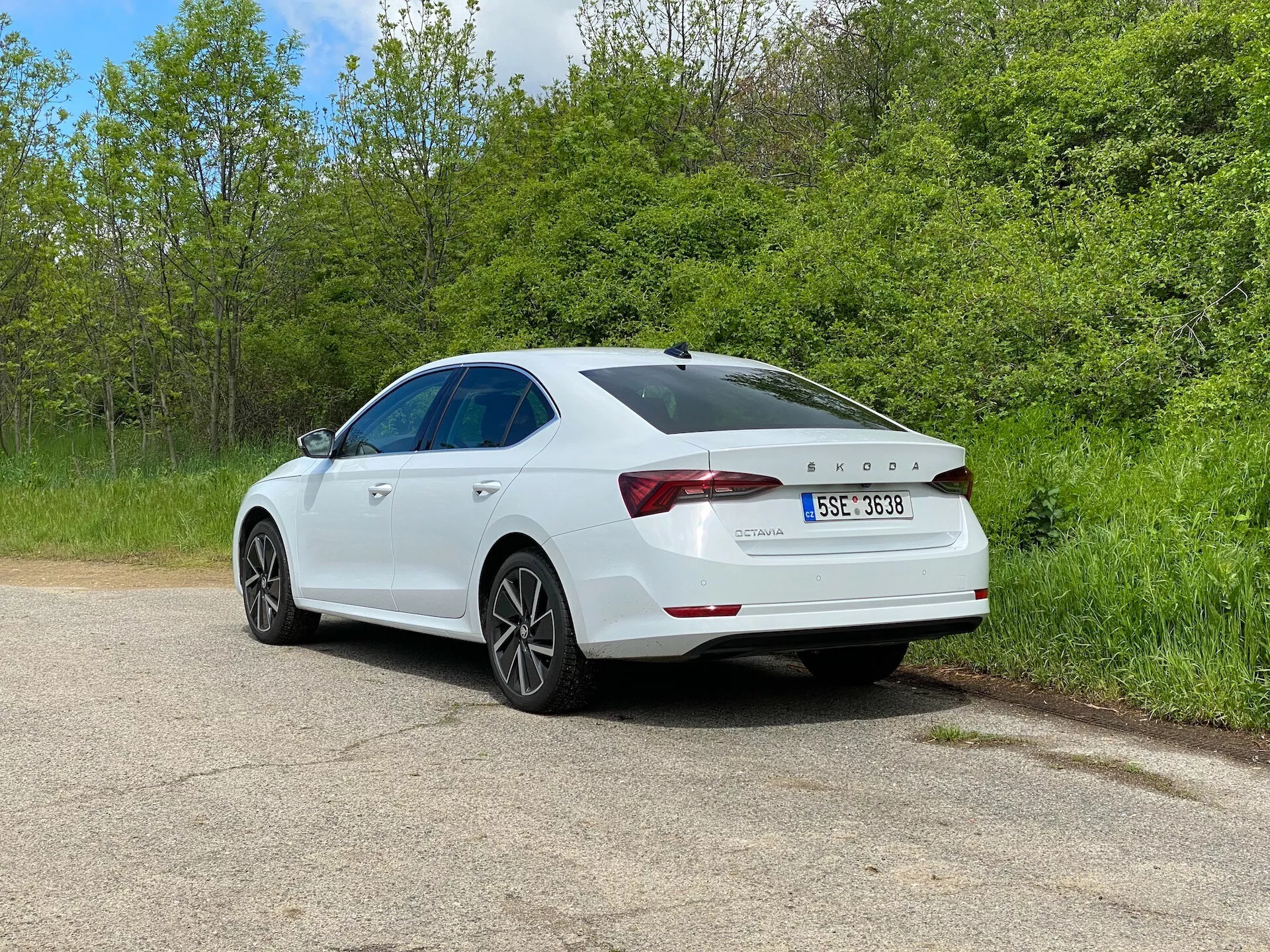 Biały sedan Skoda Octavia 2020 w trzy-czwartej tylnej perspektywie zaparkowany na żwirowej drodze, otoczony zielenią, z widokiem na charakterystyczne tylne światła, spojler tylny, felgi aluminiowe i widoczny szyberdach, podkreślający nowoczesny design i technologiczne cechy, w tym czujnik parkowania na zderzaku.