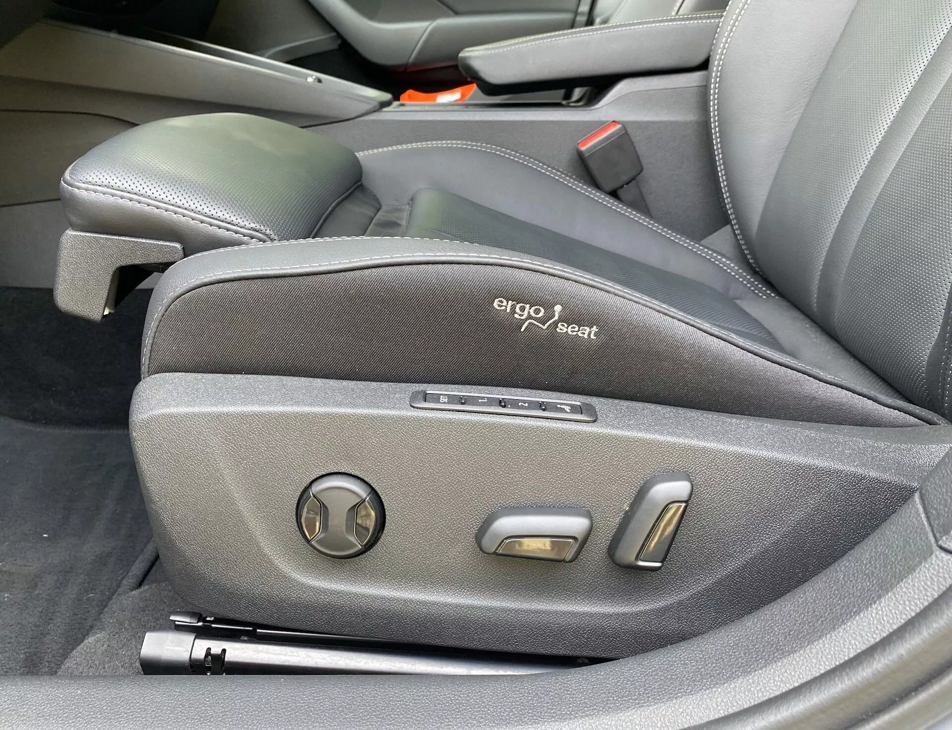 Zbliżenie bocznych kontrolek fotela kierowcy w Skodzie Octavii 2020, przedstawiające ergonomiczny design z ciemnym tapicerstwem, widoczną odznaką 'ergo seat' oraz elektronicznymi kontrolkami regulacji, w tym okrągłym regulatorem nachylenia i dwoma metalowymi przyciskami do pozycjonowania, podkreślającymi premium i technologicznie zaawansowane wnętrze pojazdu.