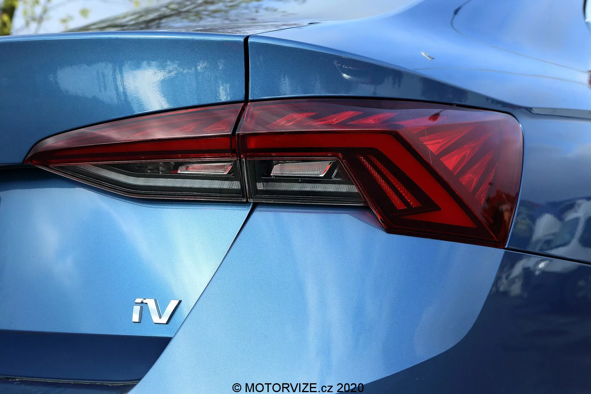 Zbliżenie na tylną część Škody Octavia Limousine 2020, skupione głównie na lewym zespole tylnych świateł, widzianym pod kątem trzech czwartych. Nowoczesny design tylnych świateł charakteryzuje się ostrymi kątami i wyrazistymi elementami LED, które są płynnie zintegrowane z liniami nadwozia samochodu. Widoczny odznaka 'iV' na błyszczącym metalicznym niebieskim bagażniku sugeruje pojazd hybrydowy lub elektryczny.