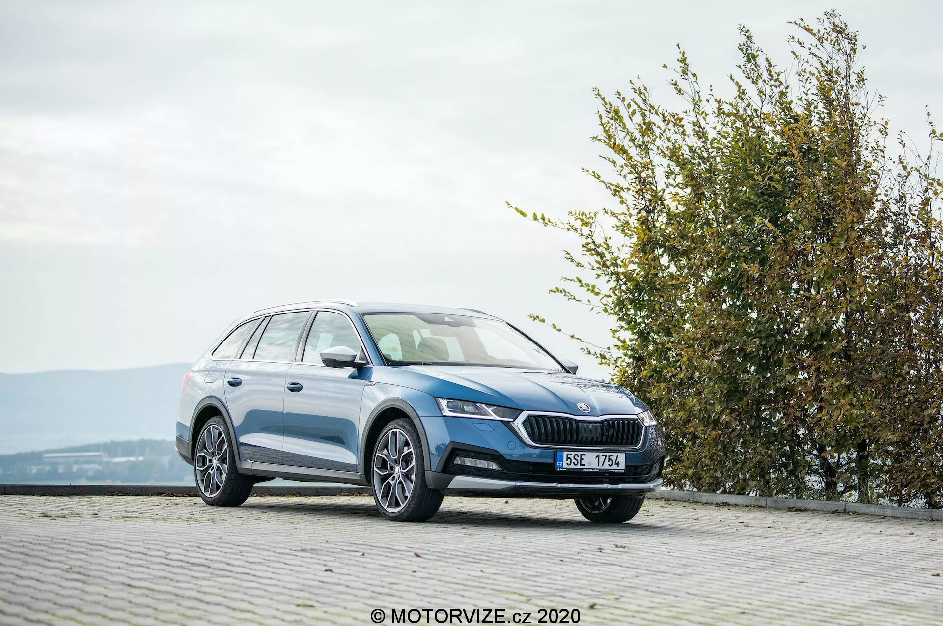 Duży kombi Skoda Octavia Scout 2020, sfotografowany z trzy czwarte przedniego widoku na brukowanym terenie na podwyższeniu. Obraz ukazuje przedni grill z wyraźnym emblematem, eleganckie, kątowe reflektory, zintegrowane światła przeciwmgielne, gładki boczny profil z aluminiowymi felgami, relingi dachowe i błyszczące wykończenie lakieru. Otoczenie obejmuje żywopłot lub krzew po prawej stronie, sugerując bliskość parku lub zielonej przestrzeni, w sprzyjających warunkach pogodowych.