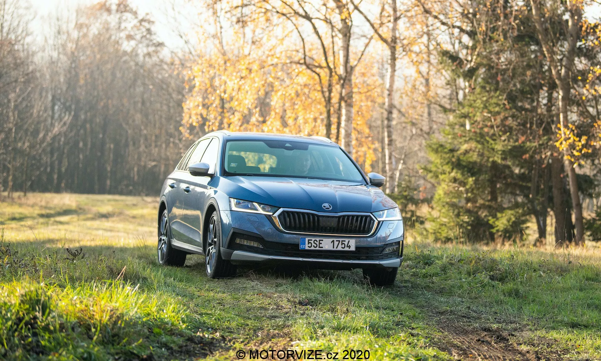 Niebieski sedan Skoda Octavia Scout 2020, sfotografowany z przodu pod kątem trzech czwartych, zaparkowany na trawiastym terenie z lasem w tle. Obraz ukazuje nowoczesne reflektory LED, charakterystyczną przednią kratkę, światła przeciwmgielne, wieloramienne felgi oraz zintegrowane kierunkowskazy w lusterkach bocznych. Ustawienie wydaje się wcześnie rano lub późnym popołudniem z miękkim światłem, nadającym ciepły ton czystemu samochodowi przeznaczonemu do reklamy lub recenzji. Rozmyte tło podkreśla cechy designu pojazdu i jego atrakcyjne otoczenie na świeżym powietrzu.