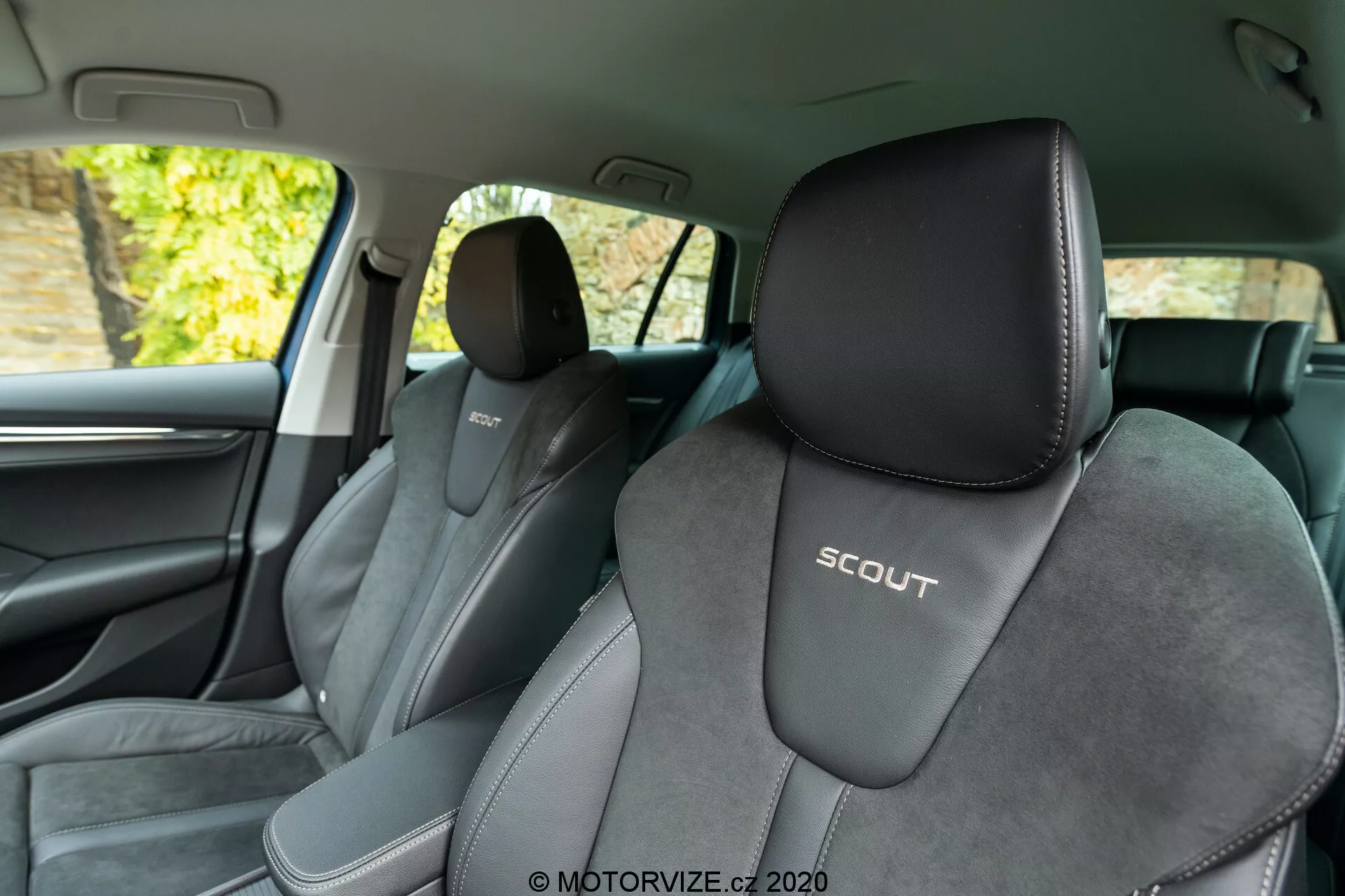 Widok wnętrza Skoda Octavia Scout 2020 z tylnego siedzenia, ukazujący przednie siedzenia. Kąt faworyzuje lewą stronę, co daje wyraźny widok na fotel kierowcy z wytłoczonym logo 'SCOUT'. Fotele są tapicerowane w dwóch odcieniach, głównie czarnym z jaśniejszymi szarymi akcentami oraz środkowym materiałem przypominającym zamsz, podkreślającym sportowy design. Obraz ukazuje również słupek B, ciemne wykończenia wnętrza, fragment przednich drzwi, kratkę głośnika i chromowaną klamkę. Tło za oknami wydaje się przedstawiać liście, co sugeruje, że samochód jest zaparkowany na zewnątrz przy naturalnym świetle.