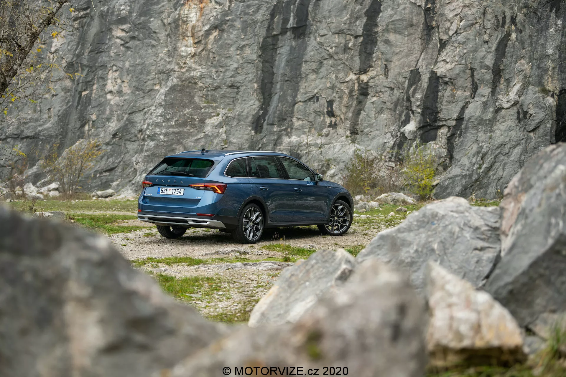 Zdjęcie przedstawia samochód Skoda Octavia Scout 2020 zaparkowany w skalistym terenie, z kamieniołomem lub pionową ścianą skalną w tle, uchwycony z lekkiej perspektywy teleobiektywu. Kąt trzy czwarte z tyłu ukazuje szczegółowy widok bocznego profilu i tyłu pojazdu, prezentując nowoczesny design kombi, opadającą tylną linię dachu i ciemnoniebieski lakier. Widoczne elementy to tylne światła, kształt okien, tylna wycieraczka, wzór kół i lusterka boczne. Otoczenie nawiązuje do tematów przygody i eksploracji, sugerując, że samochód nadaje się do pokonywania trudnego terenu i miejsc trudno dostępnych.