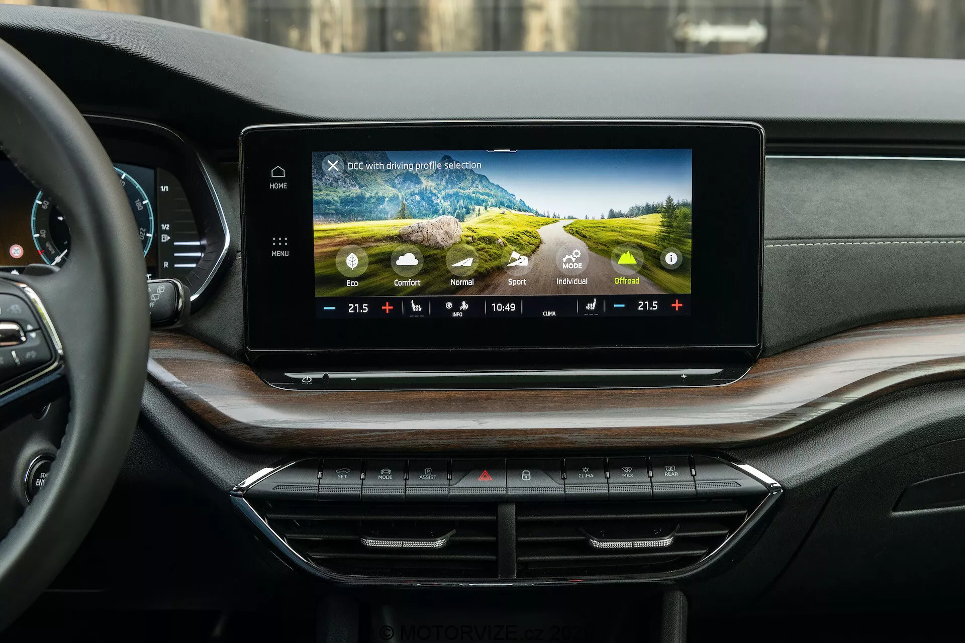 Widok wnętrza Skody Octavia 2020 (Octavia Scout) prezentujący nowoczesny system informacyjno-rozrywkowy i deskę rozdzielczą, sfotografowany z perspektywy tylnego przedziału pasażerskiego. Szeroki, poziomo zorientowany ekran dotykowy na desce rozdzielczej wyświetla kolorowe grafiki, ustawienia klimatyzacji, czas i temperaturę zewnętrzną. Poniżej znajdują się starannie uporządkowane przyciski i pokrętła kontrolne klimatyzacji. Luksusowa deska rozdzielcza ma miękkie w dotyku materiały i fornir drewniany, z dyskretnie zintegrowanymi wlotami powietrza.