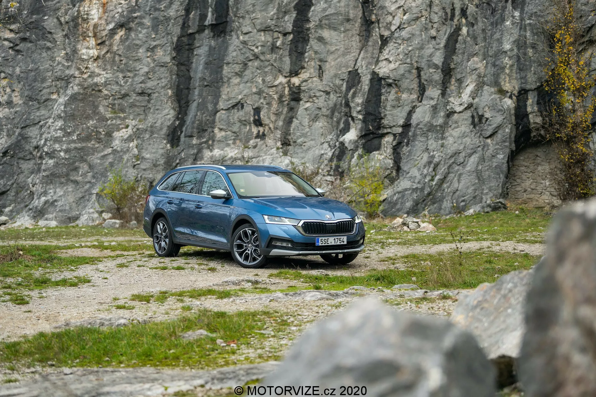Widok boczno-przedni niebieskiego kombi Skoda Octavia Scout 2020 na żwirowym terenie z skalistym tłem, pokazujący nowoczesne reflektory LED, wyrazisty przedni grill, aluminiowe felgi, lusterka boczne, obramowania okien, relingi dachowe i czyste metaliczne wykończenie, sugerując zaawansowane funkcje takie jak technologia oświetlenia, monitorowanie ciśnienia w oponach i możliwe systemy bezpieczeństwa.
