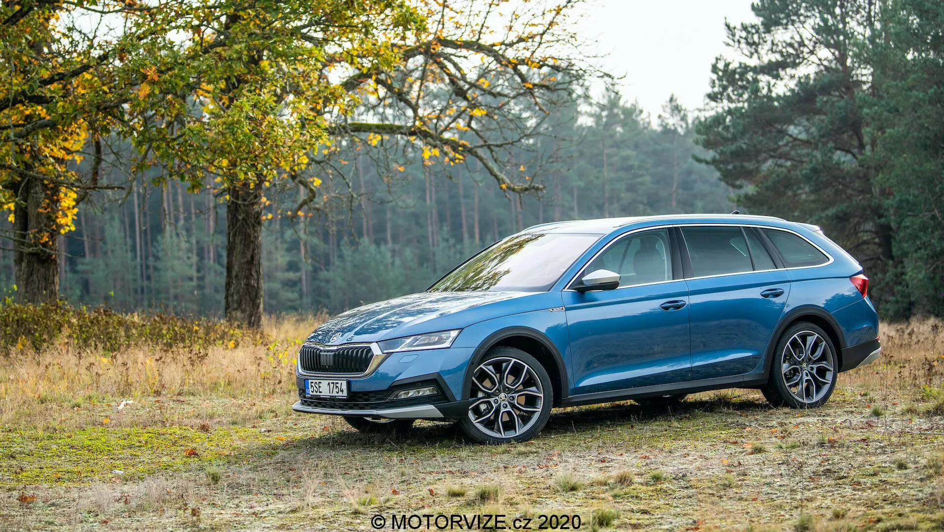 Profil boczny niebieskiego kombi Skoda Octavia Scout 2020 na drodze gruntowej z tłem drzew i trawy, pokazując jego zdolność radzenia sobie w terenie. Samochód ma metaliczny lakier, wieloramienny wzór felg aluminiowych, reflektory LED, charakterystyczną przednią kratę, relingi dachowe, lusterka boczne i klamki w kolorze nadwozia oraz dach panoramiczny. Elementy designu obejmują linię postaci biegnącą od przedniego nadkola do tylnych świateł oraz otaczające tylne światła.