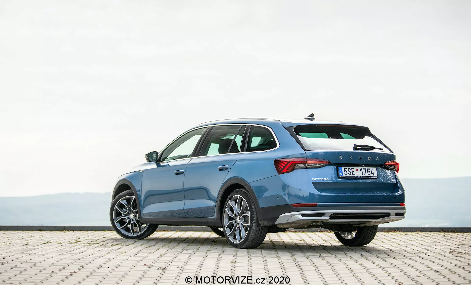 Szczegółowe zdjęcie niebieskiego kombi Skoda Octavia Scout z 2020 roku, zaparkowanego na powierzchni z płytek, uchwycone z perspektywy trzy czwarte od tyłu. Fotografia podkreśla elegancki profil pojazdu, charakterystyczny design tylnej pokrywy i prawą stronę. Widoczne cechy to poziome światła tylne, linia charakteru sugerująca ruch, tylny zderzak z odblaskami prawdopodobnie kryjącymi czujniki parkowania, podwójny układ wydechowy oraz aluminiowe felgi wieloramienne. Obraz sugeruje przestronną kabinę, zaawansowane funkcje bezpieczeństwa oraz nowoczesne elementy designu idealne dla entuzjastów technologii motoryzacyjnych.