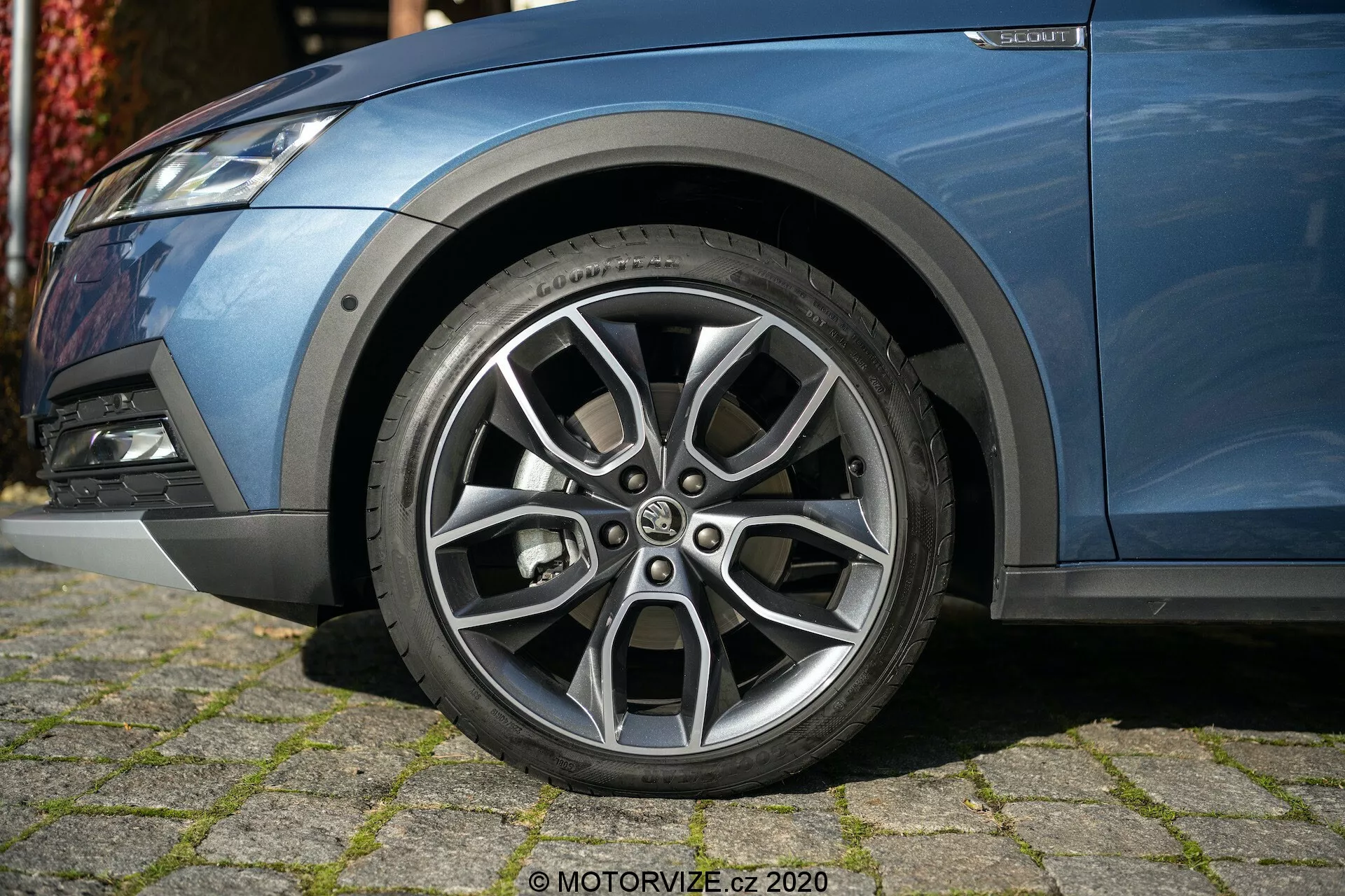 Zbliżenie lewej przedniej strony Skoda Octavia 2020 (Octavia-Scout), skupiające się na precyzyjnie zaprojektowanym kole ze stopu metali lekkich z wieloszprychowym wzorem, kontrastującym srebrnym wykończeniem i widocznymi wzorami bieżnika. Zdjęcie, wykonane pod niskim kątem, ukazuje część niebieskiej metalicznej karoserii, przedni zderzak z lampą przeciwmgielną i komponenty zawieszenia, prezentując nowoczesny, sportowy wygląd.