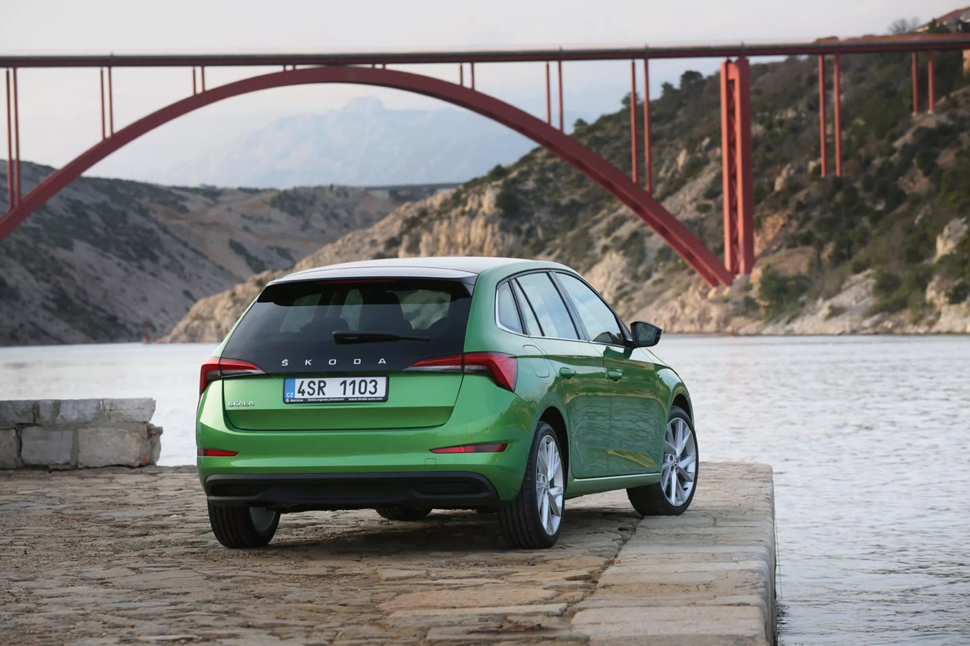 Zielona Skoda Scala z 2019 roku zaparkowana nad wodą z widokiem tyłu i części boku pojazdu, prezentująca tylne światła, klapę bagażnika, logo i oznaczenie modelu na tle czerwonego mostu i gór