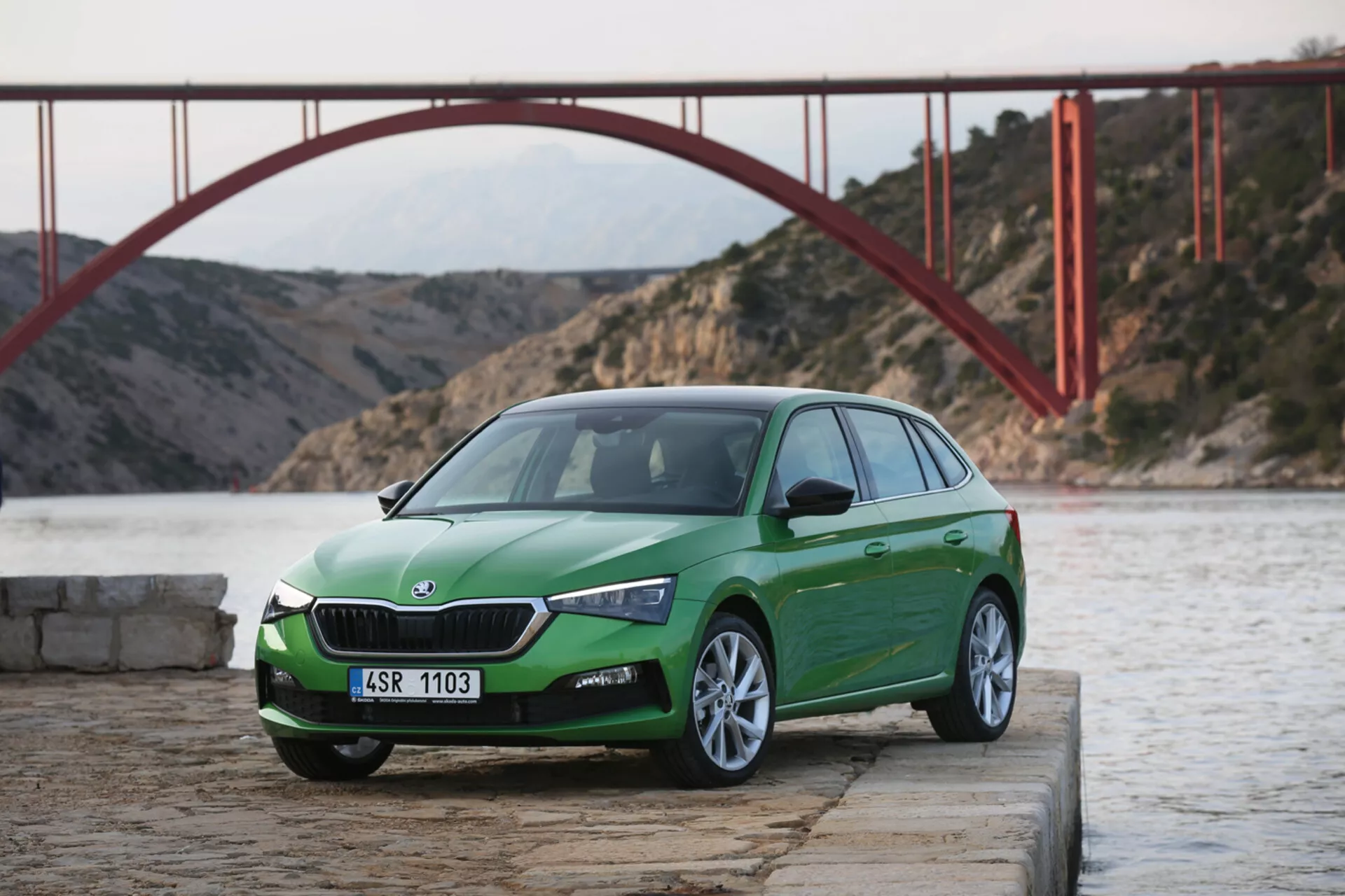 Ukośny przedni trzy-czwarty widok zielonego hatchbacka Skoda Scala 2019 na betonie przy wodzie z czerwonym mostem w tle w świetle dziennym, pokazujący design i funkcje