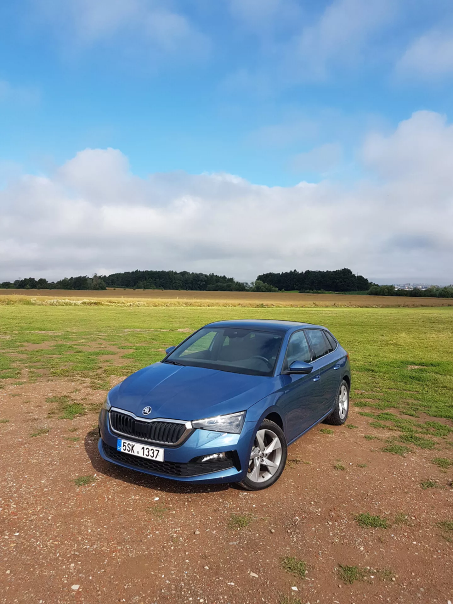 Sedan Skoda Scala rocznik 2019 w kolorze niebieskim, zaparkowany na trawie pod pochmurnym niebem, prezentujący przedni grill, reflektory i profil strony pasażerskiej na tle krajobrazu wiejskiego