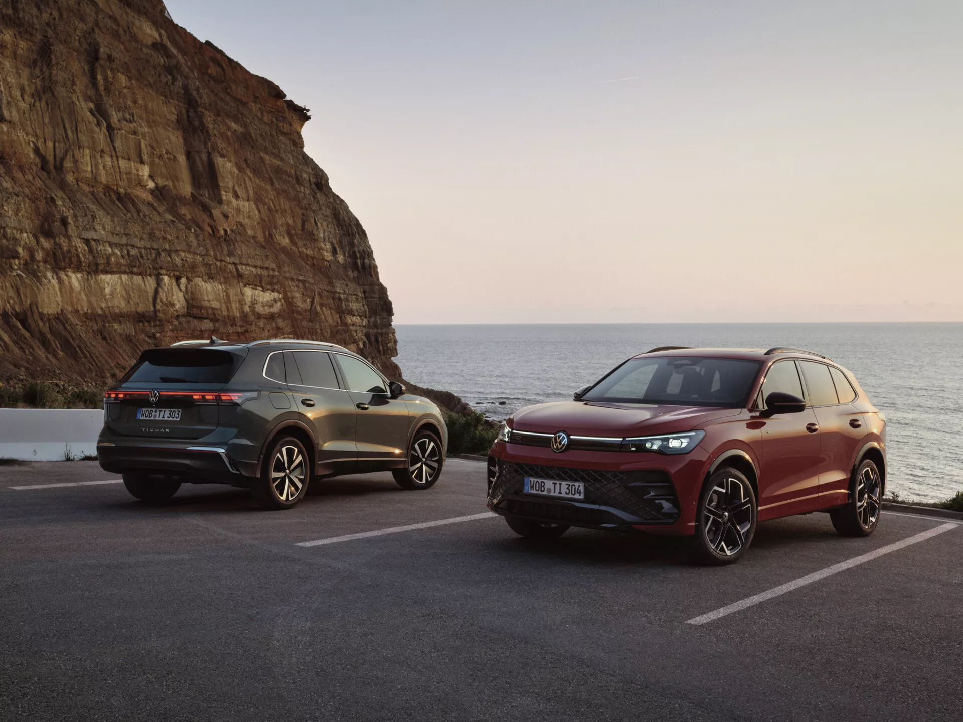 Luksusowe SUV-y w malowniczej nadmorskiej scenerii o zachodzie słońca, prezentujące model w kolorze szarym z tylnymi światłami LED i sportowymi elementami designu oraz model w kolorze bordowym z grillą przednią o orientacji sportowej i zaawansowanymi systemami wspomagania kierowcy