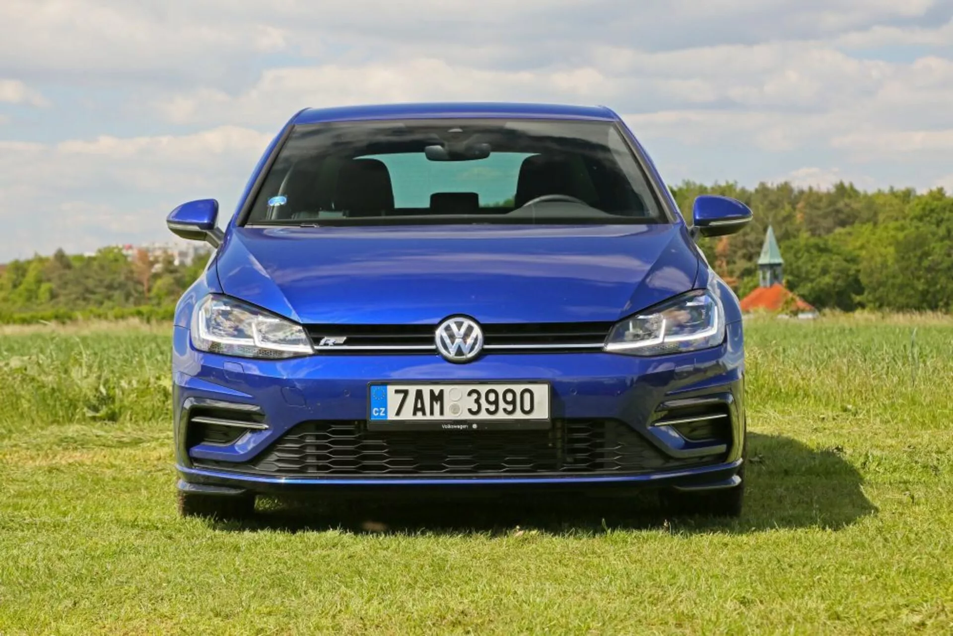 Przedni widok niebieskiego Volkswagena Golfa zaparkowanego na trawiastym polu pod bezchmurnym niebem, podkreślający jego metaliczne wykończenie, design grilla, technologię oświetlenia LED i sportowy przedni zderzak w tym wloty powietrza i emblemat.