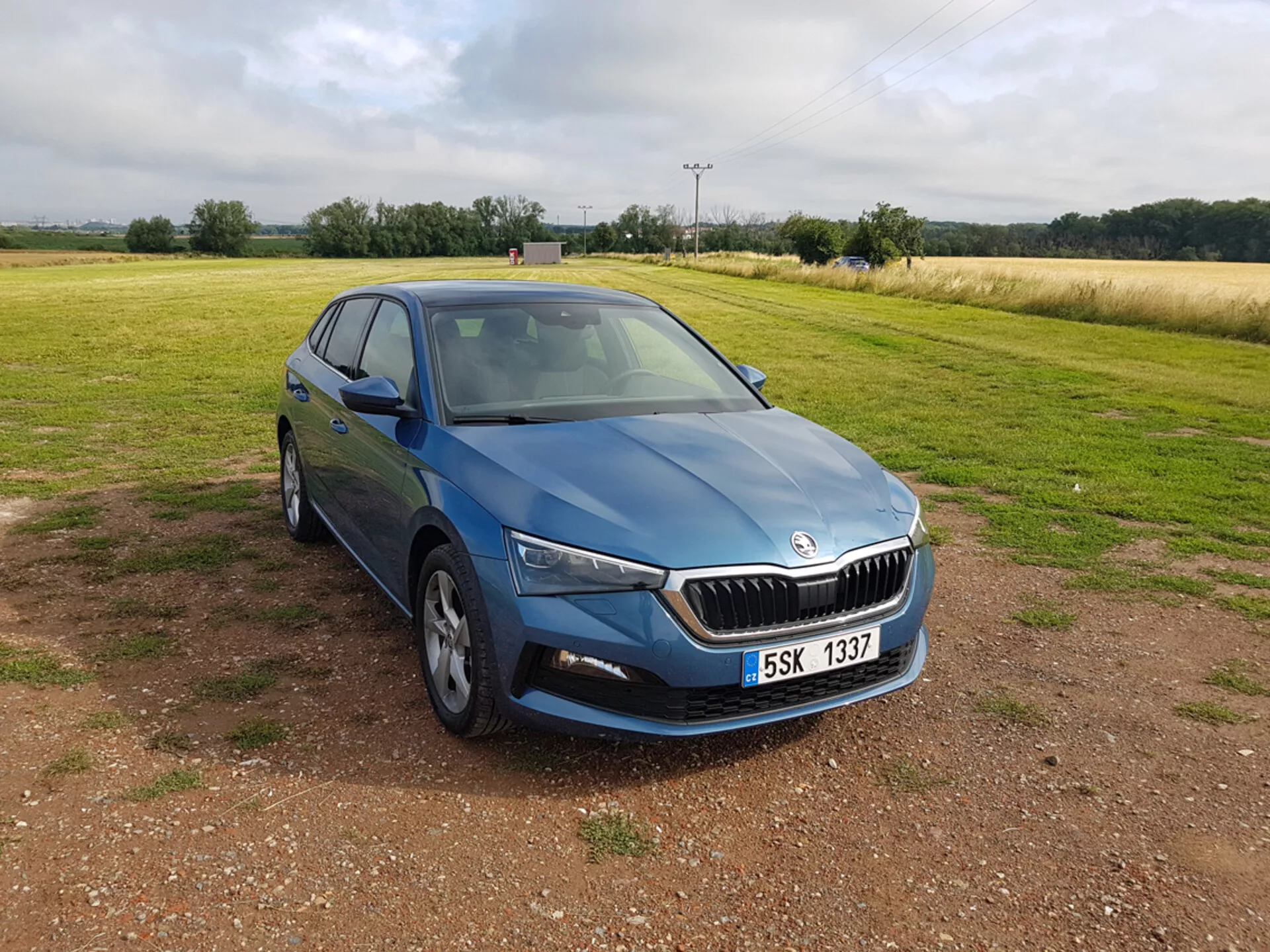 Niebieska Skoda Scala 2019 widziana pod kątem trzy czwarte z przodu, podkreślająca design, reflektory LED, chromowaną kratę, felgi aluminiowe o wielu szprychach, kierunkowskazy zintegrowane z lusterkami bocznymi i przyciemniane szyby, zaparkowana na żwirze z polem w tle pod zachmurzonym niebem.