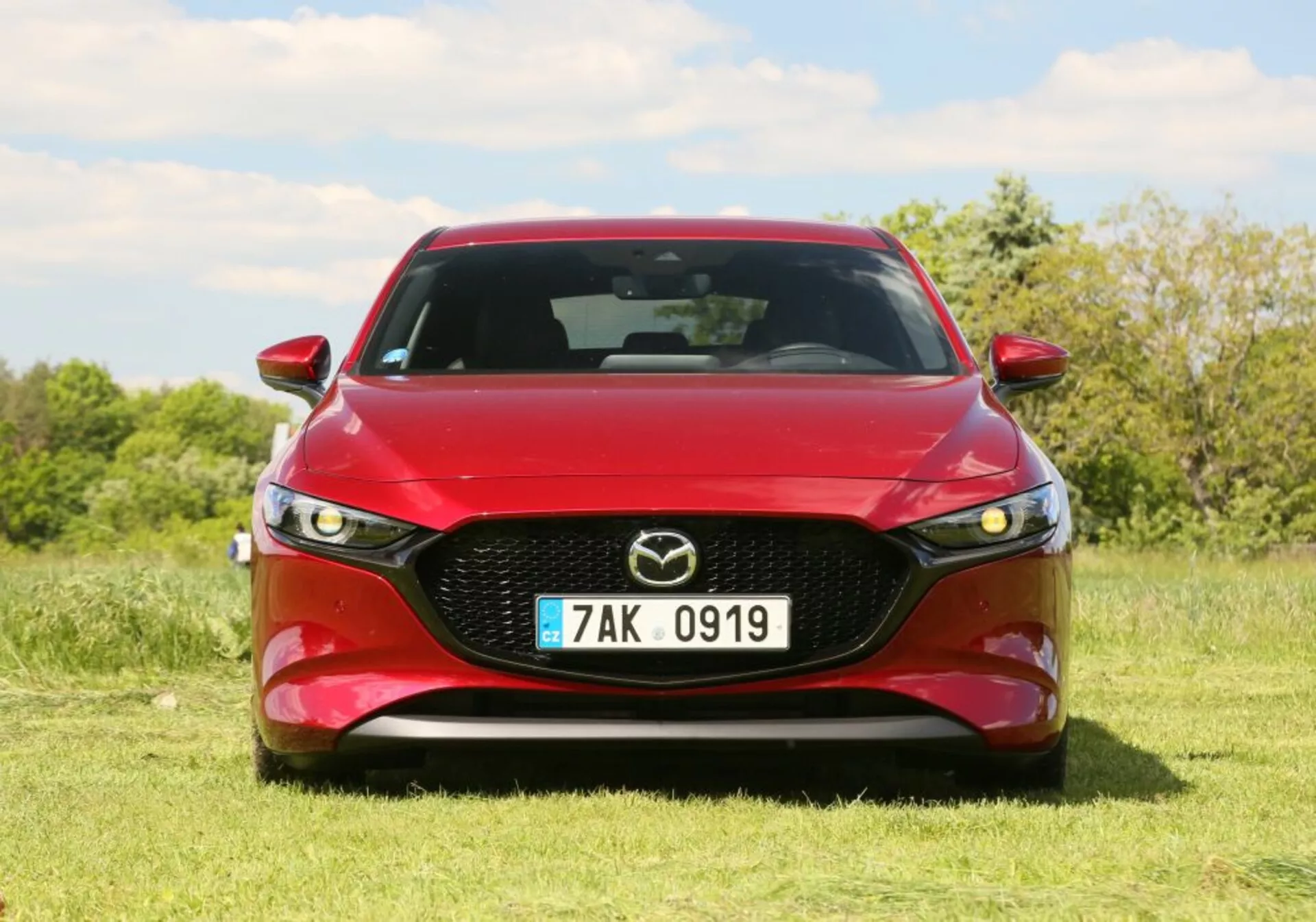 Samochód Mazda 2019 w kolorze czerwonym, prezentujący widok z przodu z wyraźną kratką z emblematem Mazda, reflektorami LED i profilowanym designem maski. Umieszczony na zewnątrz na trawie z drzewami i niebieskim niebem w tle.