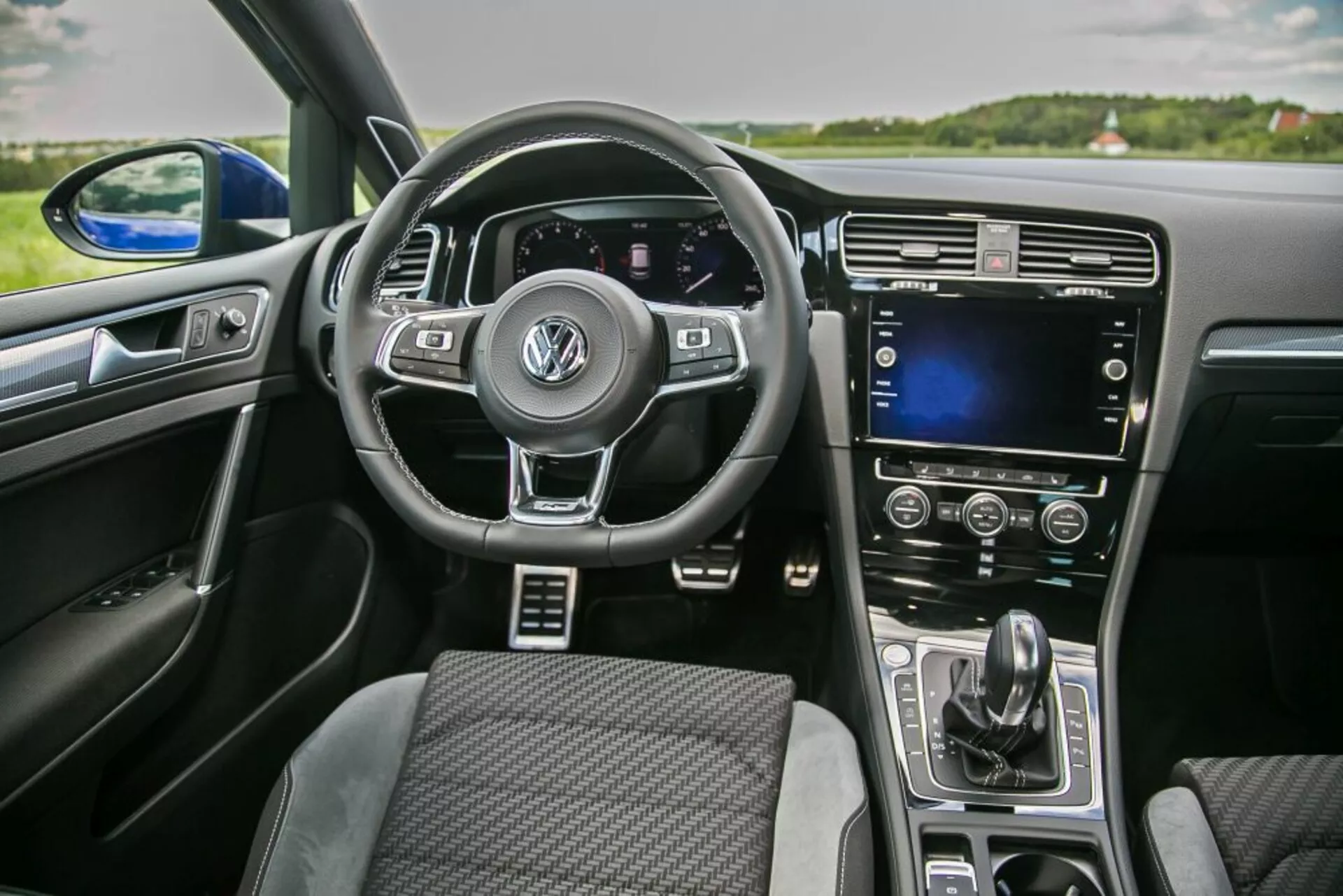Widok wnętrza Skoda Scala 2019 z perspektywy przedniego pasażera przedstawiający wielofunkcyjną kierownicę z logo Volkswagen, cyfrowy panel instrumentów, dotykowy ekran na konsoli środkowej i automatyczną kontrolę klimatu