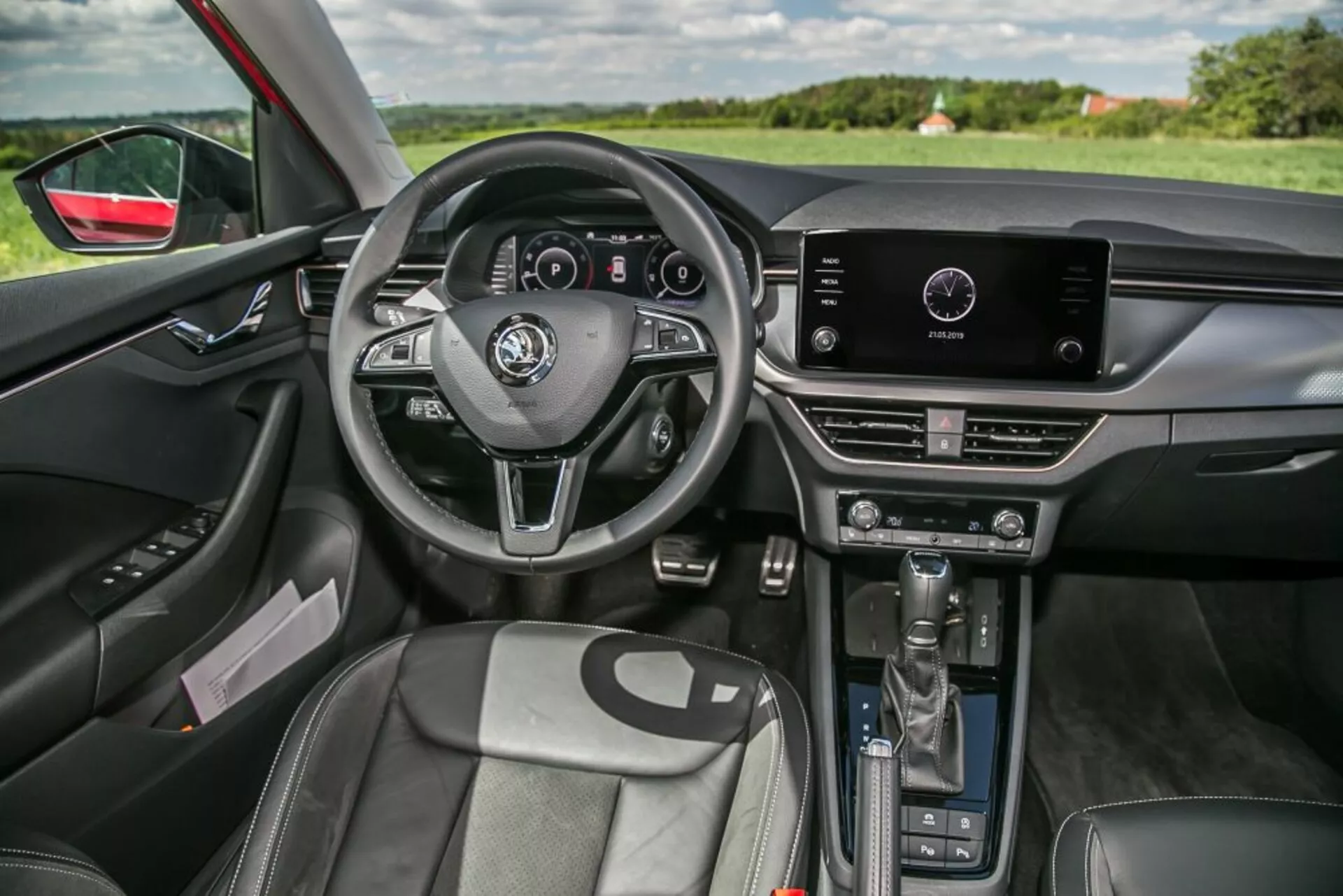 Wnętrze Skoda Scala 2019 z perspektywy pasażera przedniego z trójramienną kierownicą z przyciskami i cyfrowym zestawem wskaźników, systemem infotainment na dotyk na górnej części konsoli środkowej z manualną kontrolą klimatyzacji poniżej, automatyczną dźwignią zmiany biegów, przednimi siedzeniami z podparciami i tapicerką przypominającą skórę, panelem drzwi kierowcy z elektrycznymi regulacjami okien i lusterek, wewnętrznym oświetleniem ambientowym, lusterkiem wstecznym z funkcją przyciemniania, oraz widocznością krajobrazu przez przednią szybę i okno po stronie kierowcy, podkreślając ergonomię projektu i zintegrowaną technologię.