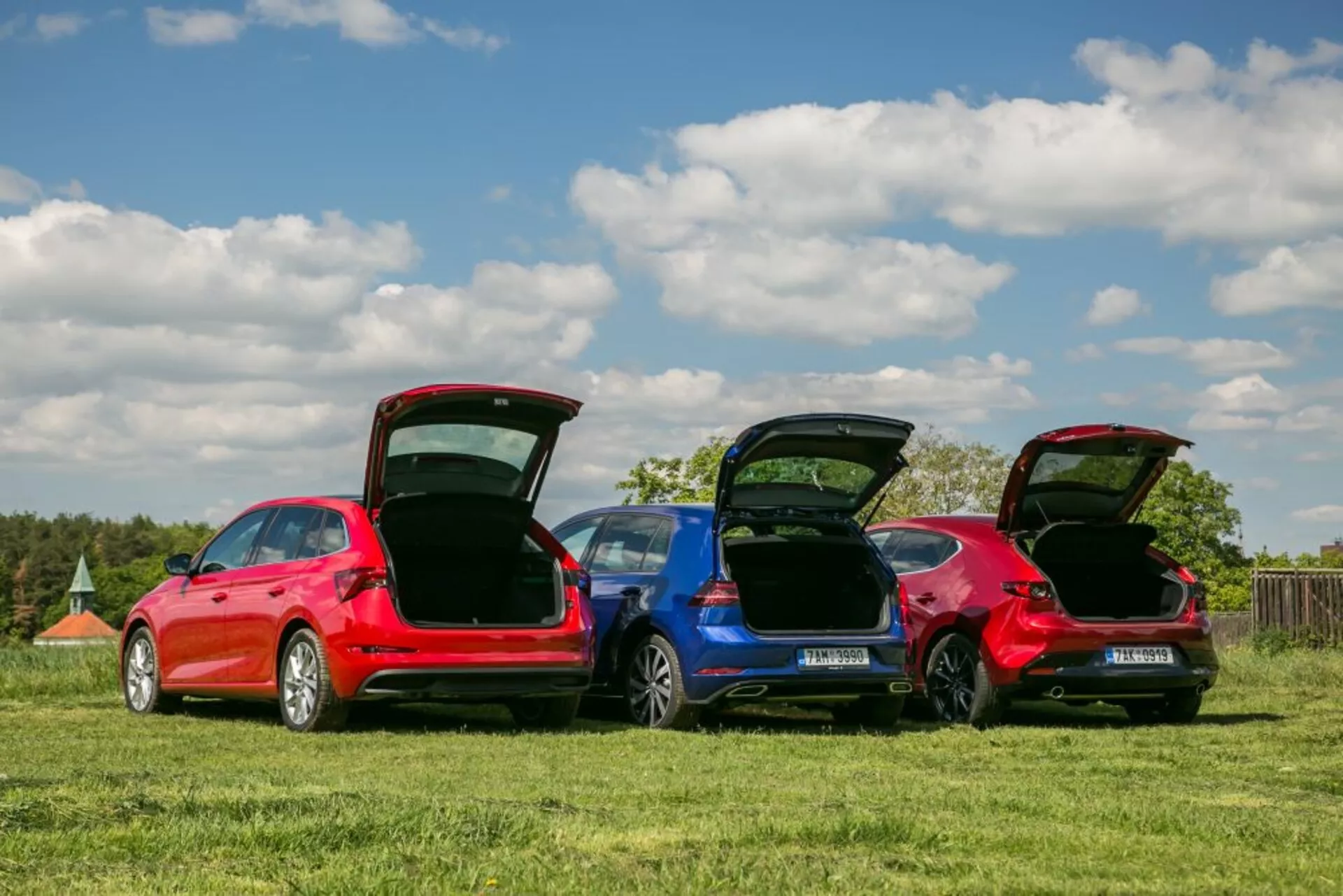Modele Skoda Scala 2019 w kolorach czerwonym i niebieskim zaparkowane na trawiastym polu, prezentujące tylne profile, otwarte bagażniki do funkcjonalności przestrzeni ładunkowej i szczegóły konstrukcji pod jasnym niebem.