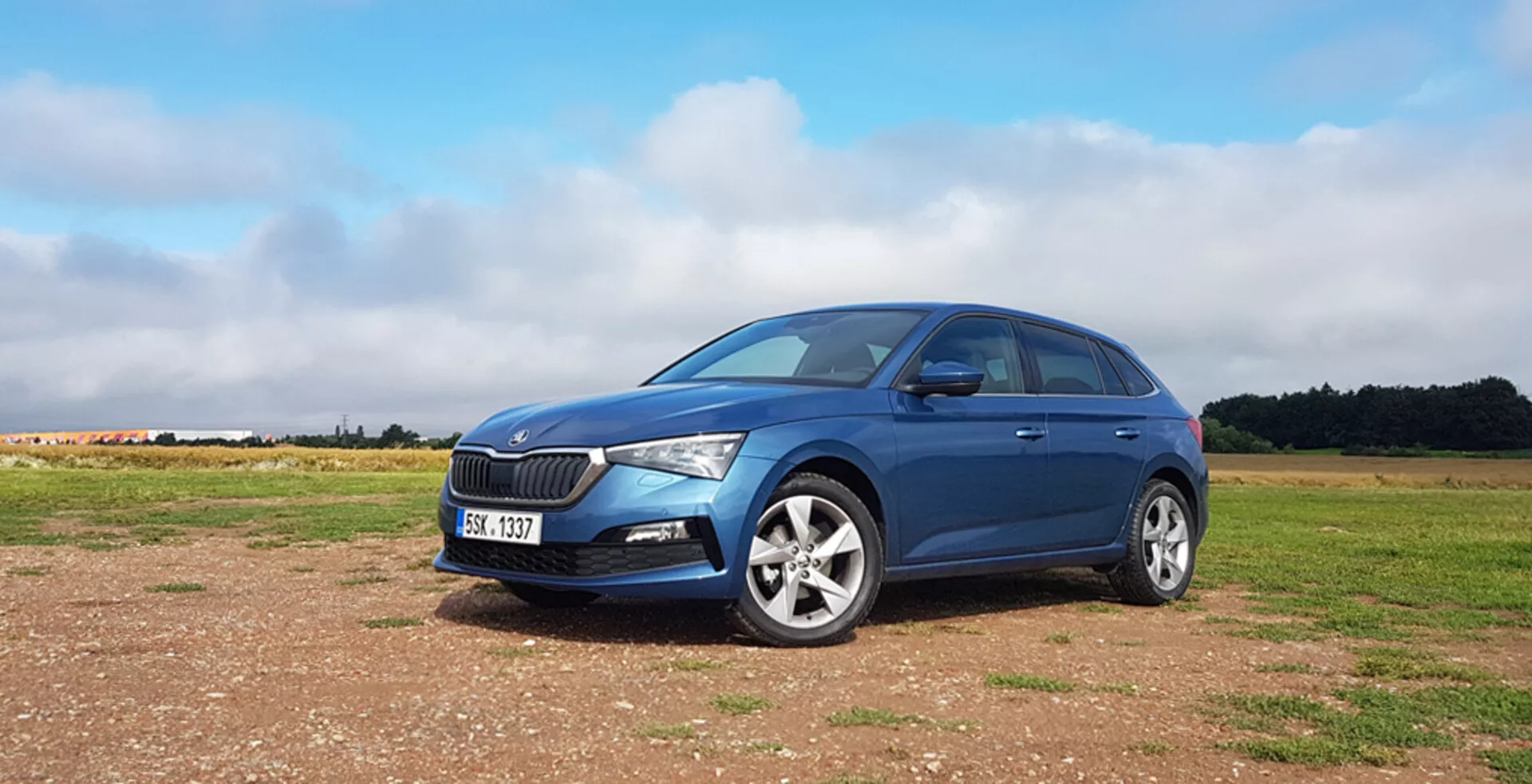 Niebieski hatchback Skoda Scala z 2019 roku na nieutwardzonej nawierzchni z charakterystyczną przednią kratką, nowoczesnymi reflektorami LED, obręczami ze stopu i eleganckim profilem bocznym strony kierowcy na tle częściowo zachmurzonego nieba