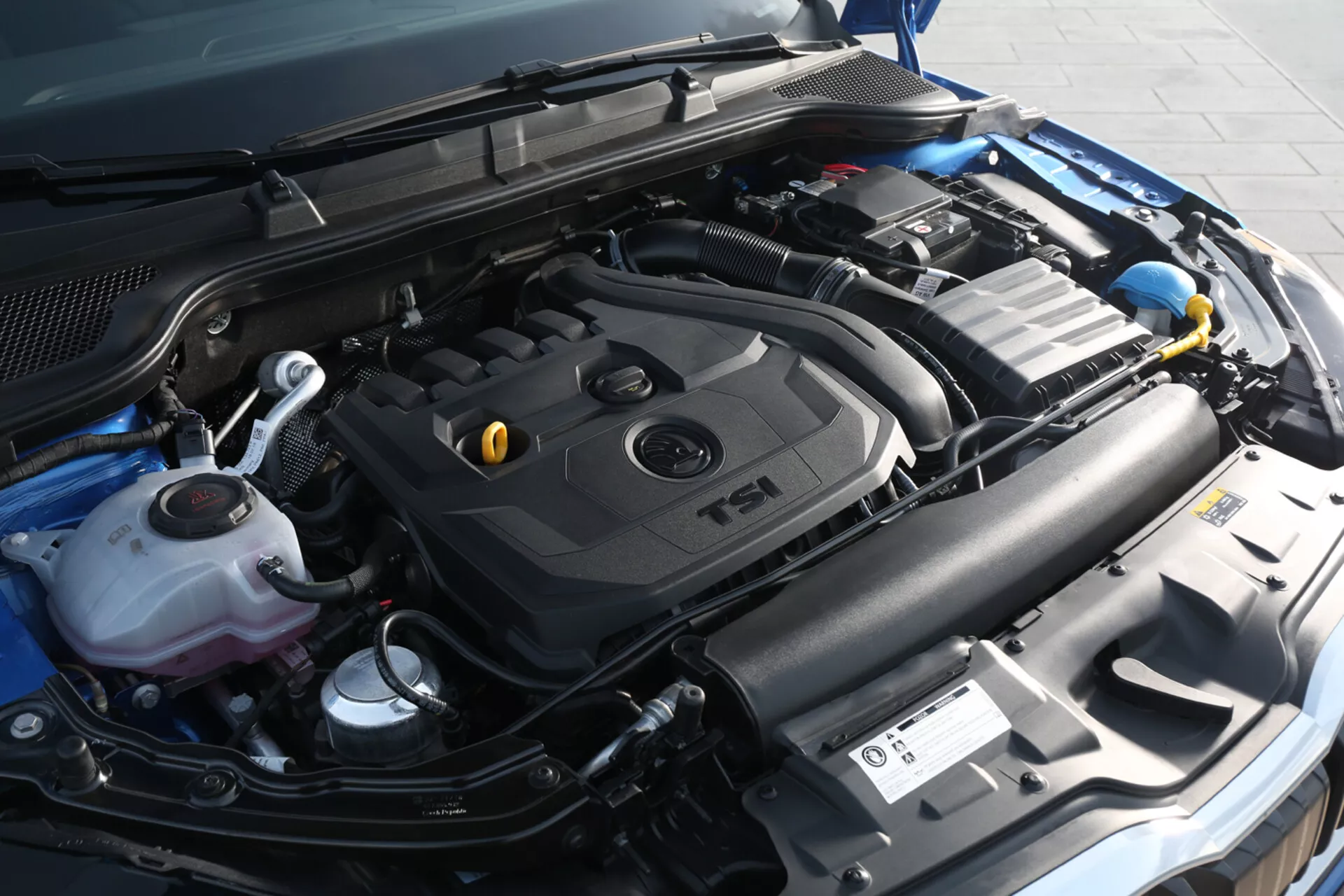 Zbliżony widok na komorę silnika Skoda Scala 2019, pokazujący pokrywę silnika TSI z turbodoładowaniem i komponenty w tym zbiornik płynu chłodzącego, pojemnik na płyn do spryskiwaczy, baterię samochodową i system dolotowy z lekko prawego przedniego kąta