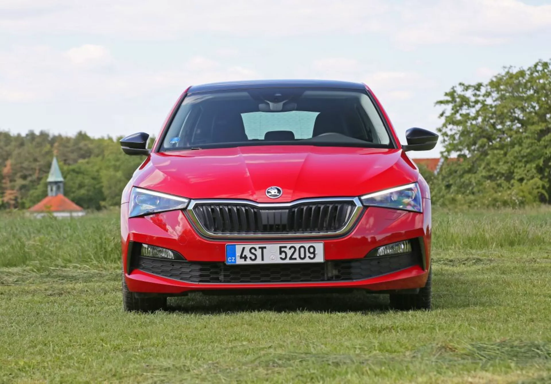 Czerwona Skoda Scala 2019 usytuowana na trawiastym polu, widok z przodu, prezentująca przedni grill samochodu, ostre reflektory i logo Skody, na naturalnym tle z drzewami i nutką niebieskiego nieba.