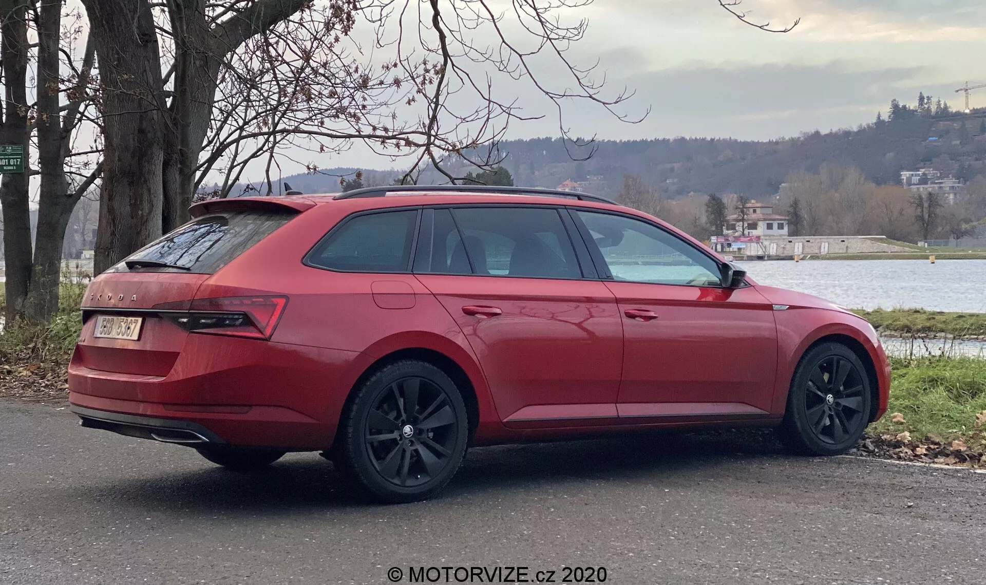 Czerwony kombi Skoda Superb 2019 zaparkowany na zewnątrz, widziany pod kątem tylno-bocznym, pokazujący prawą stronę samochodu. Kluczowe elementy designu obejmują duże ciemne felgi aluminiowe, nachylone tylne i boczne szyby, tylne światła obejmujące bok, zamknięty tylny bagażnik z emblematem, tylny zderzak z dyfuzorem i podwójnymi wydechami, relingi dachowe oraz boczne lusterko zewnętrzne w kolorze nadwozia. Otoczenie zawiera zieleń i zbiornik wodny w tle, sugerując spokojne środowisko podmiejskie lub wiejskie.