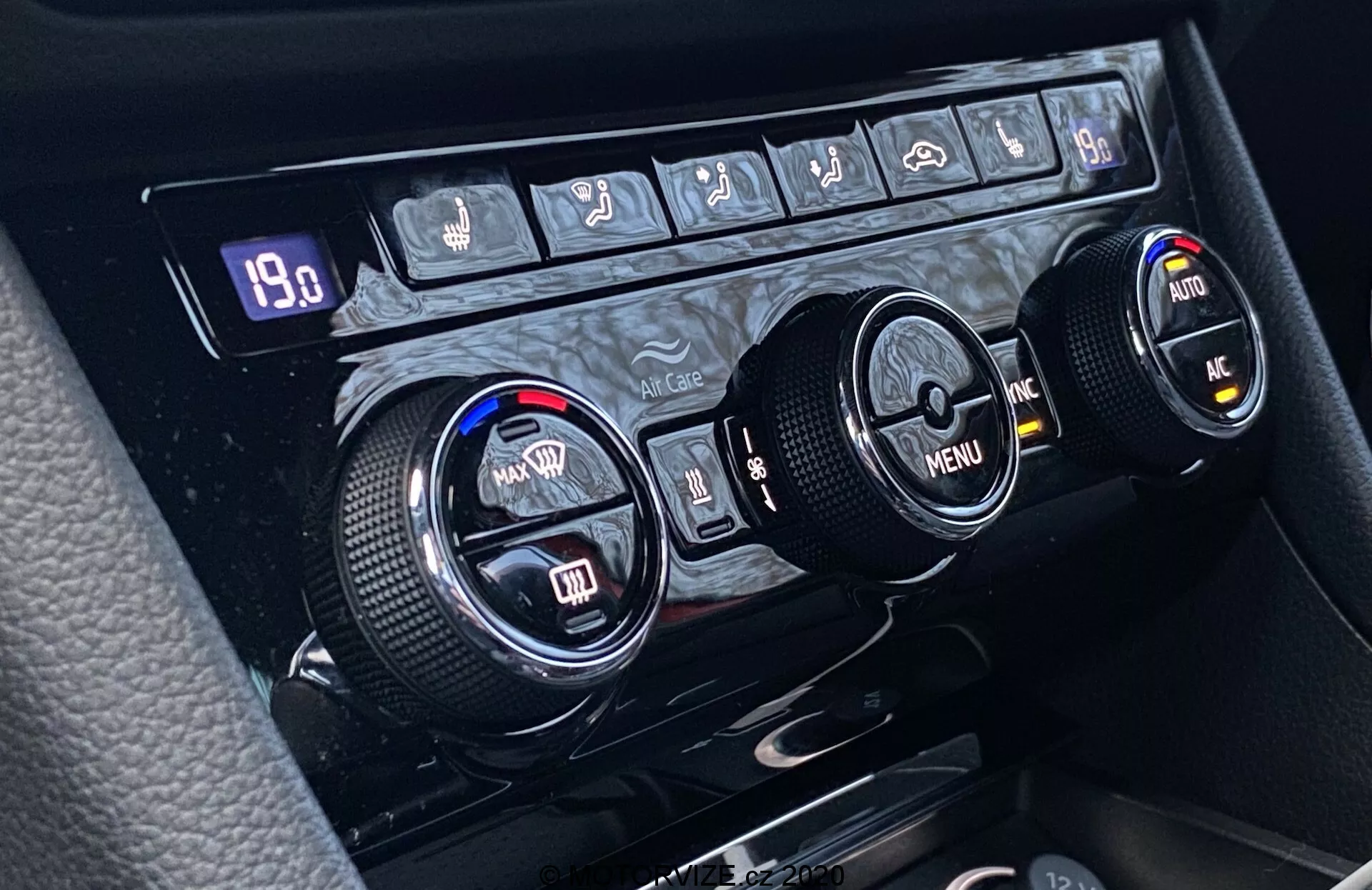Zbliżenie na system kontroli klimatu w Škoda Superb 2019 umieszczony na konsoli środkowej, z trzema okrągłymi pokrętłami do sterowania temperaturą, nawigacją po menu i dodatkowymi funkcjami. Lewe pokrętło reguluje temperaturę po stronie kierowcy, prawe po stronie pasażera, natomiast środkowe pokrętło umożliwia nawigację po menu przy użyciu strzałek kierunkowych. Teksturowane krawędzie zapewniają ergonomiczne użytkowanie. Powyżej pokręteł znajduje się rząd przycisków do sterowania rozmrażaniem tylnej szyby, odmrażaniem przedniej szyby, recyrkulacji powietrza, automatycznej klimatyzacji, klimatyzacji i możliwie przycisku awaryjnego. Wyświetlacz cyfrowy pokazuje '19.0,' co może wskazywać ustawioną temperaturę lub inną wartość związaną z działaniem systemu.