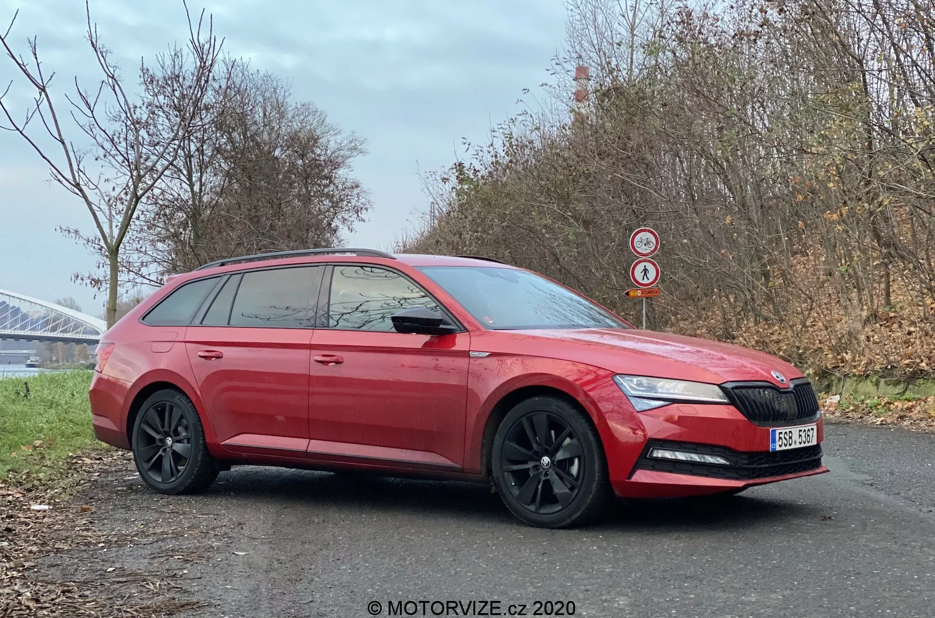 Profil boczny czerwonego kombi Skoda Superb z 2019 roku zaparkowanego przy drodze, podkreślający jego nowoczesny design z ostrymi przednimi światłami zawierającymi dzienne światła LED, charakterystycznym przednim grillem z logo, czarnymi felgami aluminiowymi, przyciemnianymi szybami zapewniającymi prywatność i wydłużoną linią dachu typową dla kombi. Zdjęcie, wykadrowane tak, aby uwydatnić zarówno bok, jak i lekko skróconą przednią część, uchwyca szczegóły takie jak zintegrowane kierunkowskazy w lusterkach bocznych, klamki w kolorze nadwozia, ochronne listwy dolne i tylny spojler dachowy. Wyraźny obraz wykonany podczas zimnej pory roku w pobliżu konstrukcji mostowej umożliwia dostrzeżenie cech samochodu i otoczenia.