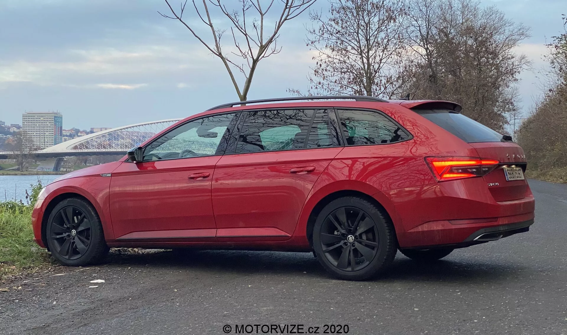 Obraz przedstawia nowoczesny, intensywnie czerwony Skoda Superb 2019 kombi widziany z kąta trzech czwartych z tyłu. Samochód zaparkowany na utwardzonym terenie z rzeką i mostem w tle, pokazuje smukłe linie, pochylony dach z tyłu, lekko przyciemniane szyby z chromowanymi wykończeniami i zintegrowany spoiler tylny. Sportowy design podkreślają duże czarne aluminiowe felgi z widocznymi zaciskami hamulców, kontrastujące czarne lusterka boczne, wpuszczane klamki drzwi, tylne światła LED oraz czujniki parkowania na tylnym zderzaku, wskazując na wysoki poziom wyposażenia z zaawansowanymi funkcjami wspomagania kierowcy.