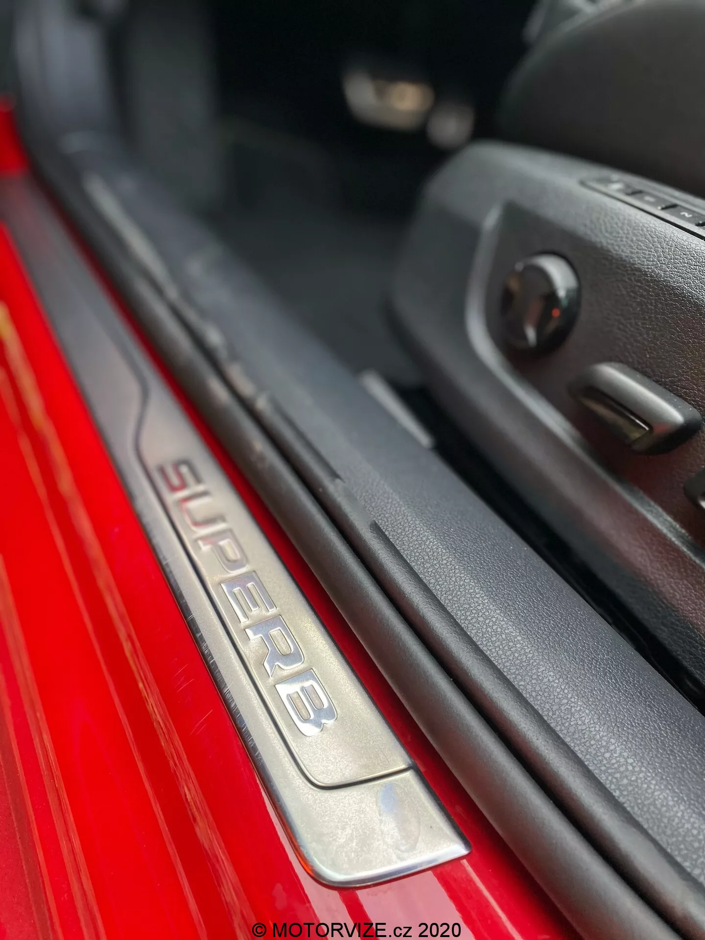 Zbliżenie na próg drzwi pasażera w modelu Škoda Superb 2019. Obraz ukazuje nachylony kąt, na którym widoczna jest inskrypcja 'SUPERB' na progu drzwi wykonanym ze szczotkowanej stali nierdzewnej, podświetlanym dla lepszego efektu estetycznego. Czerwona farba zewnętrzna wyraźnie kontrastuje z ciemnym wnętrzem. Widoczna jest krawędź drzwi z plastikowym panelem i sterowaniem szybami elektrycznymi, co podkreśla zaawansowane funkcje. Płytka głębia ostrości eksponuje próg drzwi, tworząc efekt bokeh w tle.