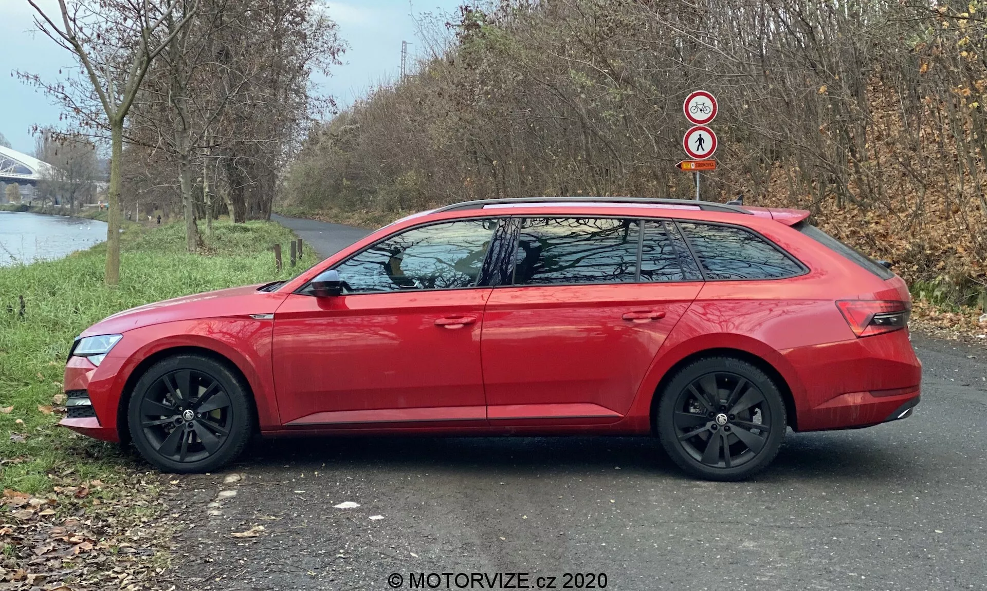Boczny profil nowoczesnego kombi Skoda Superb 2019 (Superb-Limousine) w kolorze czerwonym, pokazującego wydłużoną linię dachu, czarne felgi aluminiowe i lusterka boczne w kolorze nadwozia z wbudowanymi kierunkowskazami. Samochód jest zaparkowany na asfaltowej ścieżce obok trawnika, z drzewami i zbiornikiem wodnym w tle, oświetlony naturalnym światłem dziennym.