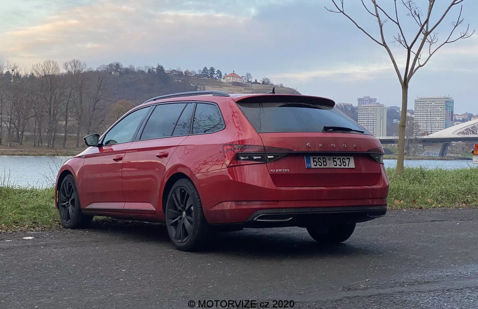 Kątny widok z tyłu czerwonego kombi Skoda Superb 2019 zaparkowanego na zewnątrz, skupiający się na tylnej i pasażerskiej stronie pojazdu. Charakteryzuje się ciągłymi tylnymi światłami na długości klapy bagażnika, podwójnymi rurami wydechowymi, przyciemnianymi szybami, felgami ze stopu o ciemnym wykończeniu i znajduje się na utwardzonej powierzchni z akwenem wodnym oraz mostem w tle.