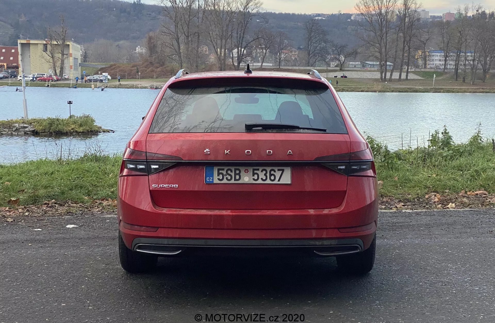 Widok z tyłu na czerwony Škoda Superb 2019 limuzyna, z charakterystycznym oznakowaniem Škoda i napisem modelu OCTAVIA na bagażniku. Zdjęcie ukazuje nowoczesne, poziome światła tylne, centralnie umieszczoną tablicę rejestracyjną, tylna wycieraczkę, przyciemnianą tylną szybę z liniami odmrażania, wysoko umieszczone światło stop, czujniki parkowania w zderzaku i odblaski. Samochód jest zaparkowany na zewnątrz z widokiem na wodę, drzewa i linię horyzontu w tle.