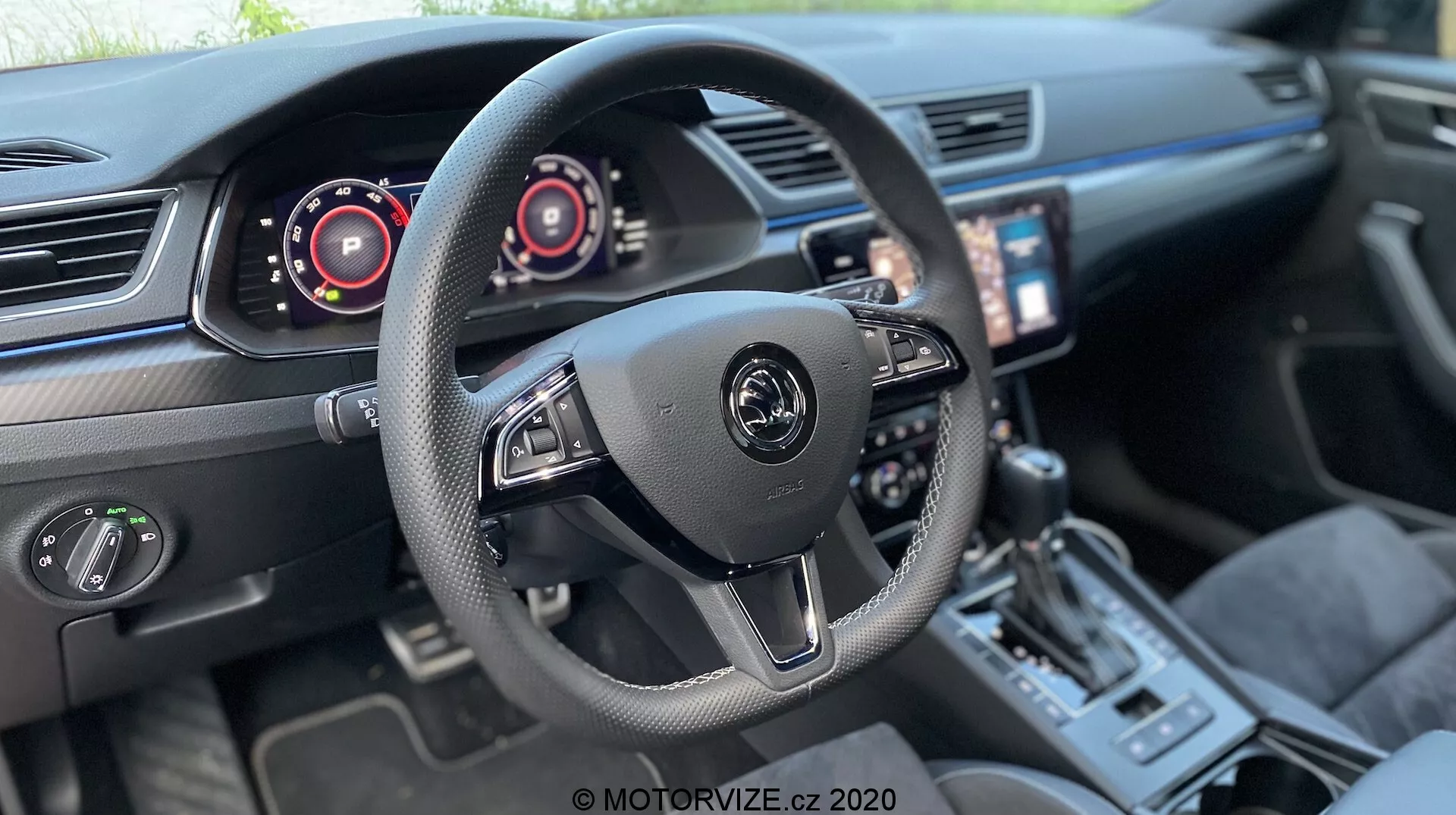Wnętrze Škody Superb Limousine 2019 skoncentrowane na obszarze kokpitu kierowcy. Centralna, trójramienna kierownica z logo producenta i zamontowanymi kontrolerami do systemu audio i systemów pokładowych, w tym łopatki zmiany biegów. Cyfrowe panele za kierownicą wskazujące prędkościomierz i obrotomierz. Ekran dotykowy systemu informacyjno-rozrywkowego skierowany w stronę kierowcy, dwustrefowa klimatyzacja, sportowa dźwignia zmiany biegów, selektor trybu jazdy oraz skórzane fotele z akcentowanymi szwami, co sugeruje nowoczesny, wysoce wyposażony pojazd z zaawansowaną technologią.