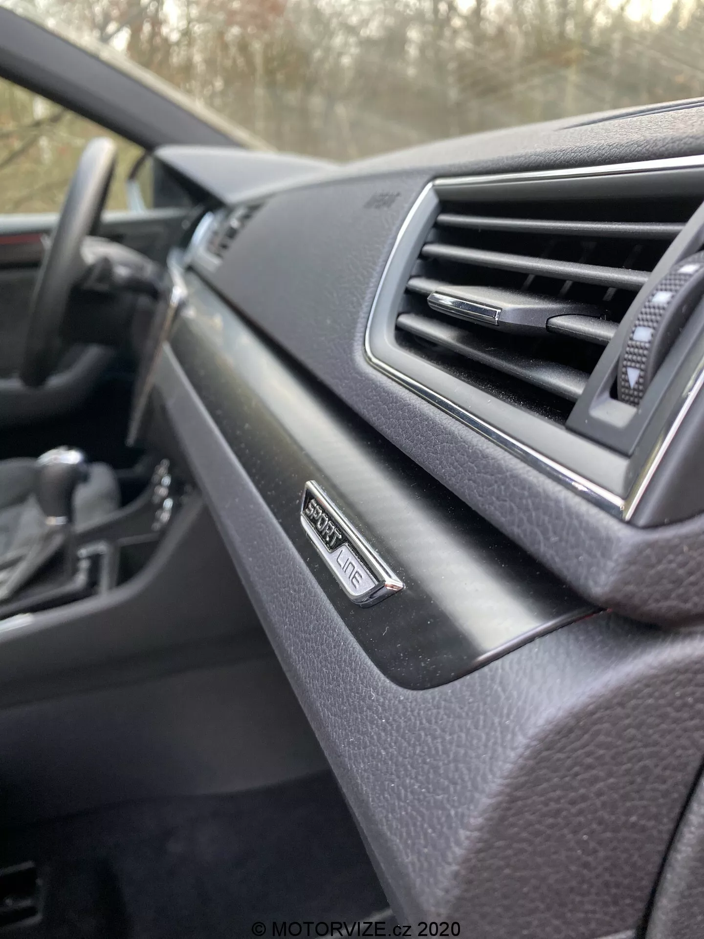 Zbliżenie wnętrza Škoda Superb 2019, deska rozdzielcza z perspektywy pasażera. Skupienie na odznace 'Superb' w pobliżu wentylacji, nowoczesny design z miękkim wykończeniem. Rozmyta kierownica w tle, ciemna kolorystyka.