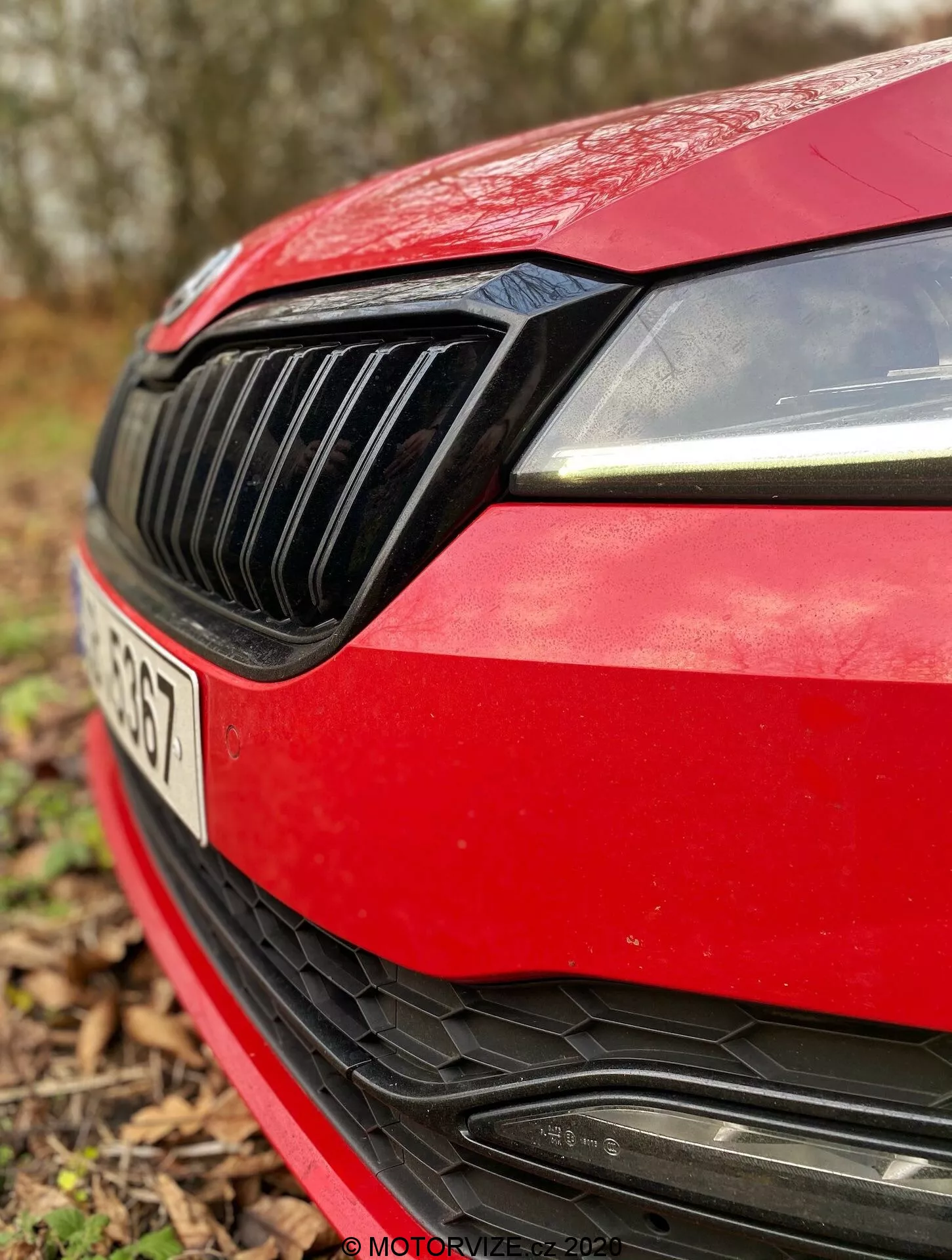 Zbliżenie przedniej lewej ćwiartki czerwonej Škody Superb Limuzynie 2019. Zdjęcie uchwyca nowoczesny design prawego reflektora z dziennymi światłami LED, czarną kratkę wlotu powietrza z pionowymi listwami, opływowy brzeg czerwonej maski, sportowy przedni zderzak z dużym wlotem powietrza z poziomymi listwami oraz miejsce na logo producenta, częściowo zasłonięte. Tablica rejestracyjna jest lekko widoczna, ale nie jest ostra ze względu na małą głębię ostrości, z rozmytym naturalnym tłem.