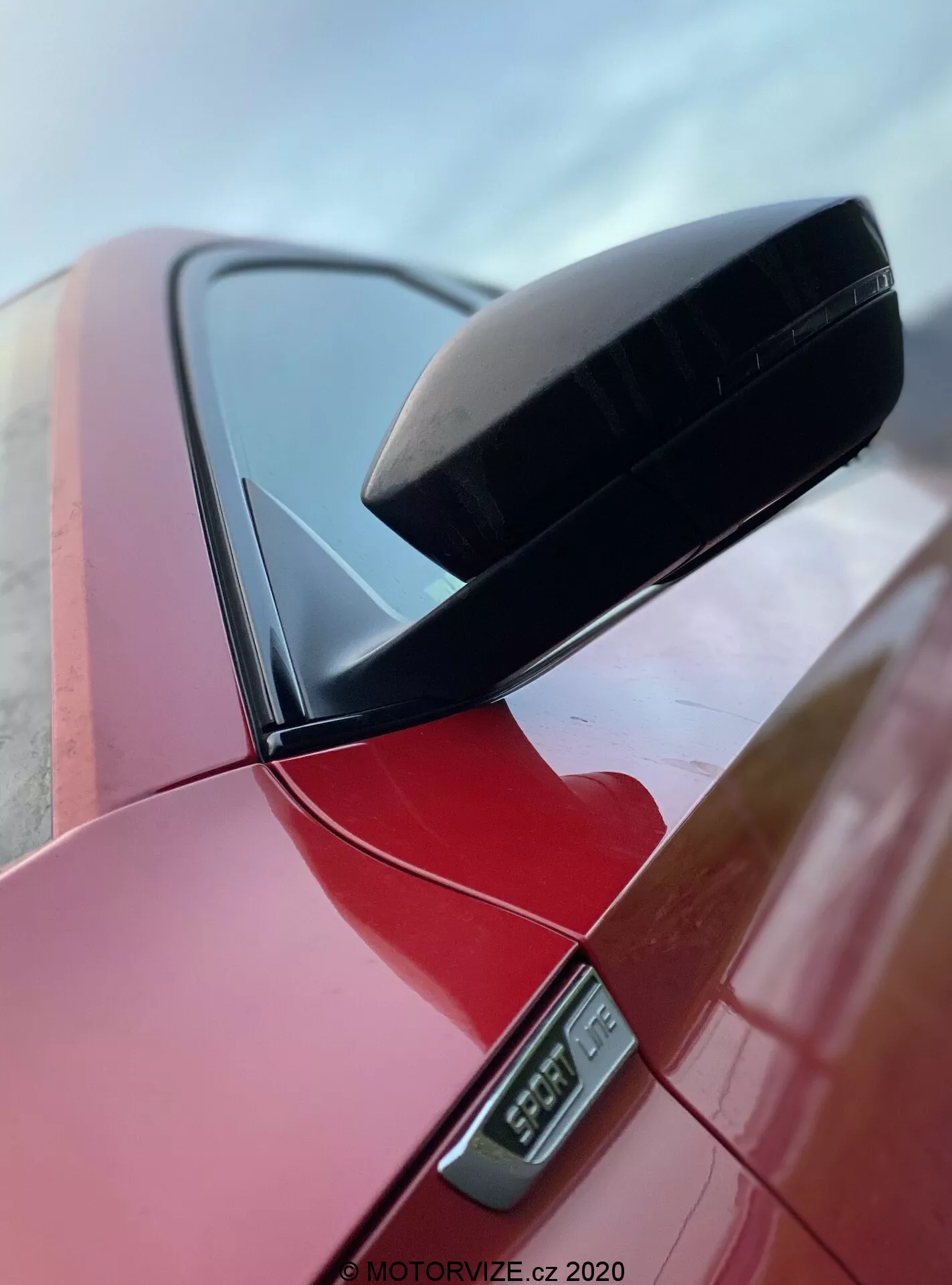 Zbliżenie lusterka bocznego w wibrująco czerwonym Skoda Superb 2019 limuzyna, podkreślające czarną aerodynamiczną obudowę lusterka i boczny wskaźnik LED pod nim, z widoczną częścią słupka samochodu, wykonane z niskiego kąta, podkreślające czysty design i wypolerowane wykończenie, oznaczone znakiem wodnym MOTORVIZE.cz 2020.