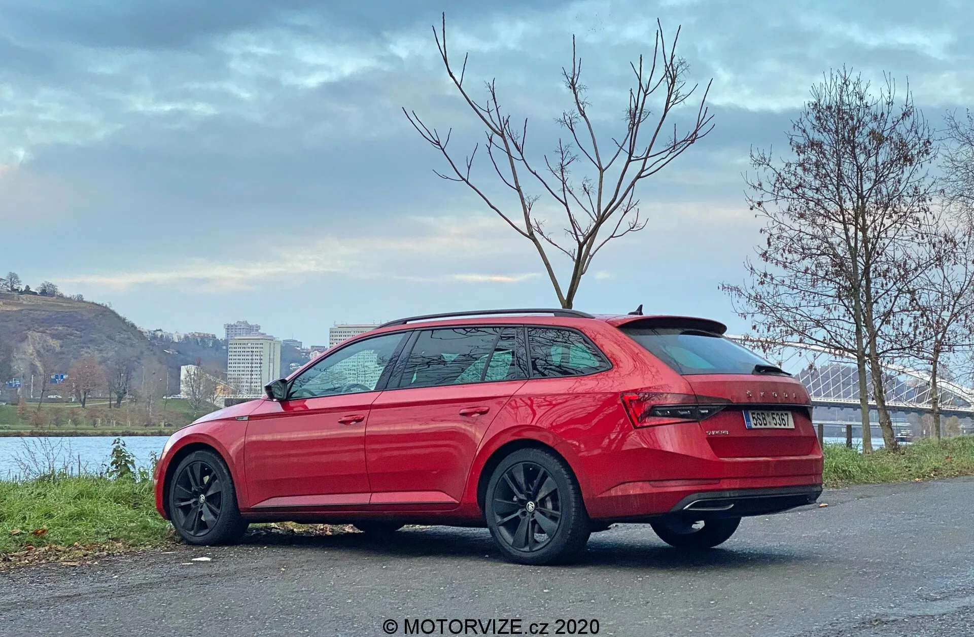Obraz Skoda Superb 2019, kombi, widok z tyłu trzech czwartych, pokazujący wysoko zamontowane tylne światła, masywny zderzak, brak widocznych rur wydechowych, spoiler dachowy, eleganckie linie boczne, klamki w kolorze nadwozia, osłony progów bocznych, chromowane listwy okien, ciemne wieloramienne koła, zaparkowany na brukowanej powierzchni w pobliżu brzegu rzeki z mostem i drzewami, zachmurzone niebo, znak wodny MOTORVIZE.cz 2020.