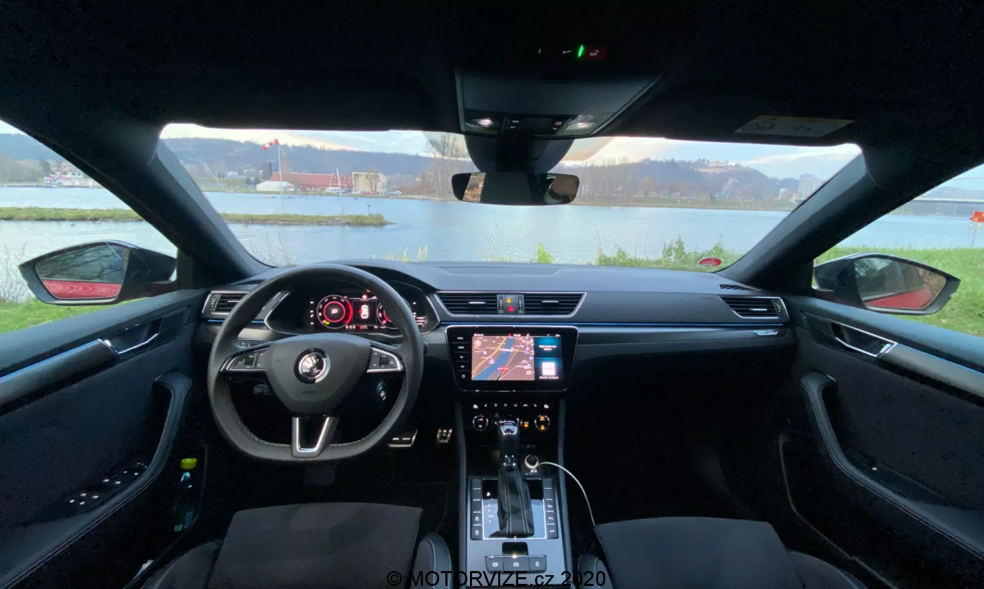 Widok wnętrza Skoda Superb 2019 z tylnego siedzenia w kierunku deski rozdzielczej, obejmujący całą szerokość wnętrza samochodu. Centralna kierownica z wbudowanymi sterownikami i logo Skoda jest wyraźnie widoczna na pierwszym planie. Za kierownicą znajduje się cyfrowa tablica wskaźników pokazująca podstawowe informacje o pojeździe. Centralna konsola zawiera system multimedialny z nawigacją i ustawieniami multimediów, a poniżej umieszczone są sterowanie klimatyzacją i przyciski funkcji. Dźwignia zmiany biegów, uchwyty na napoje oraz szczegółowo wykończone fotele z kontrastowymi szwami podkreślają nowoczesną estetykę i skupienie na komforcie oraz wygodzie kierowcy.