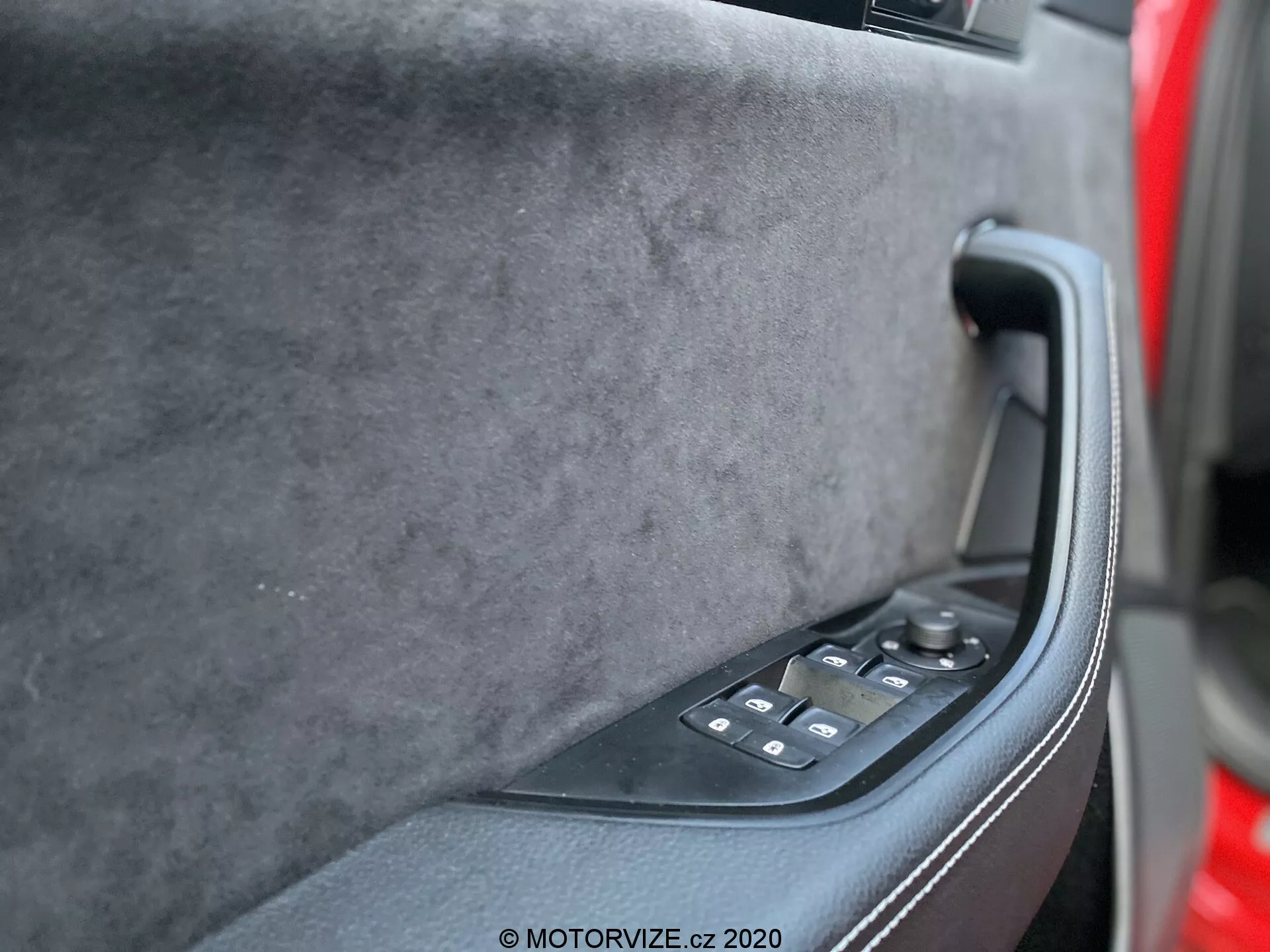 Wewnętrzny panel drzwiowy Skoda Superb 2019 z perspektywy siedzenia kierowcy, prezentujący górną część z miękkiego w dotyku materiału wysokiej jakości, solidną czarną ergonomiczną klamkę oraz błyszczący czarny panel sterowania z przyciskami regulacji elektrycznych szyb i prawdopodobnie lusterka. Stonowane ciemne kolory, nowoczesna technologia i luksusowy design są widoczne.