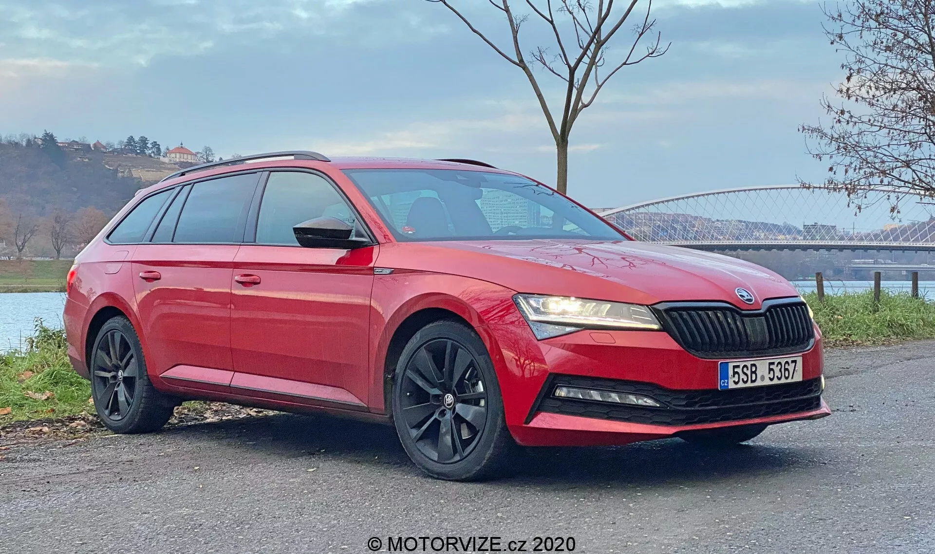 Obraz czerwonego kombi Skoda Superb 2019 uchwycony z perspektywy trzech czwartych od przodu. Zdjęcie podkreśla przedni grill z pionowymi listwami, eleganckie kątowe reflektory, światła przeciwmgielne w dolnym zderzaku, czarne wieloramienne felgi aluminiowe oraz błyszczący lakier z wyraźnymi liniami charakterystycznymi i relingami dachowymi. Widoczne są również lusterka boczne w kolorze nadwozia z wbudowanymi kierunkowskazami oraz przyciemniane tylne szyby. Pojazd, ustawiony na płaskiej powierzchni na zewnątrz, widoczny jest na tle naturalnym z mostem i zbiornikiem wodnym w oddali. Warunki dzienne podkreślają zarówno dolne, jak i górne sekcje samochodu.