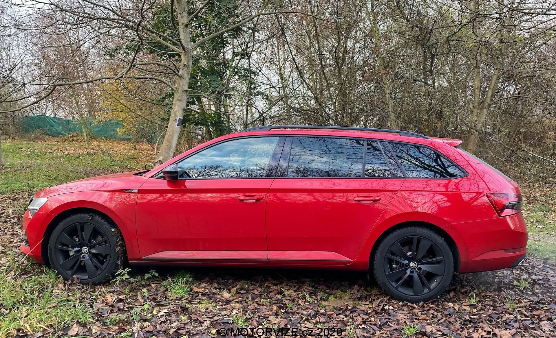 Skoda Superb kombi 2019 w jaskrawoczerwonym kolorze, ukazany z profilu bocznego. Nowoczesne cechy projektowe obejmują ostre linie geometryczne, czarne wieloszprychowe felgi aluminiowe, przyciemniane szyby z czarnymi słupkami B i pochylony dach dla eleganckiego, aerodynamicznego profilu. Samochód zaparkowany na trawiastym terenie obok ścieżki, z naturą w tle, sugerując jego dostosowanie do rodzinnych wycieczek lub przygód.