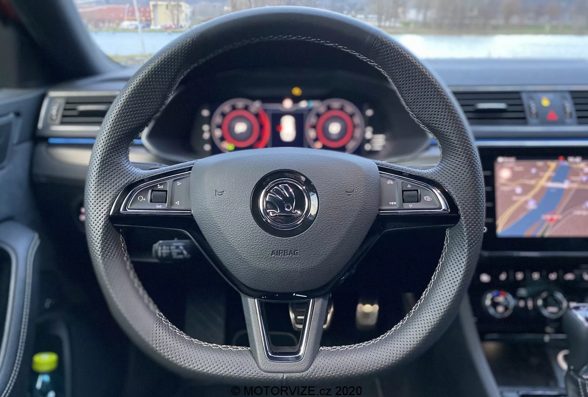 Widok wnętrza Skody Superb Limousine 2019 z perspektywy kierowcy. Siedzenie kierowcy ukazuje kierownicę owiniętą czarną skórą z przeszyciami, posiadającą różne kontrolki, w tym regulację głośności i prawdopodobne ustawienia pojazdu. W centrum obrazu znajduje się emblemat producenta z napisem 'AIRBAG', podkreślający obecność przedniej poduszki powietrznej kierowcy. Widoczne są łopatki zmiany biegów, sugerujące ręczną zmianę biegów. Zestaw wskaźników z obrotomierzem, prędkościomierzem i wyświetlaczem cyfrowym jest częściowo widoczny, podczas gdy system informacyjny pokazuje mapę nawigacji. Płytka głębia ostrości koncentruje się na kierownicy i emblematcie, a deska rozdzielcza i ekran dotykowy są lekko rozmyte.