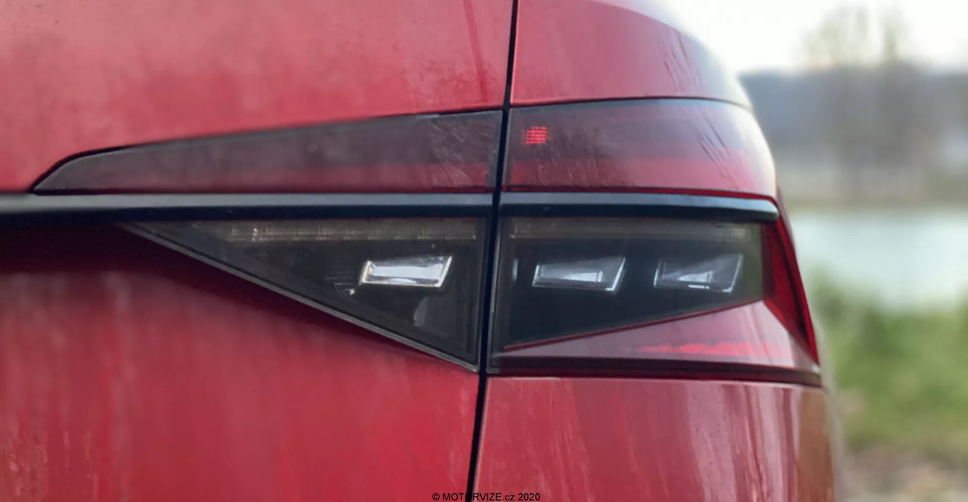 Zbliżenie tylnej części Skoda Superb 2019, skupiające się na zespole tylnych świateł pod kątem bocznym, ukazujące nowoczesny, elegancki design z poziomymi elementami LED w ciemnoczerwonej soczewce, odzwierciedlając współczesną estetykę sportową lub luksusową. Błyszczące nadwozie i czarna pozioma listwa zwiększają atrakcyjność wizualną na rozmytym tle.