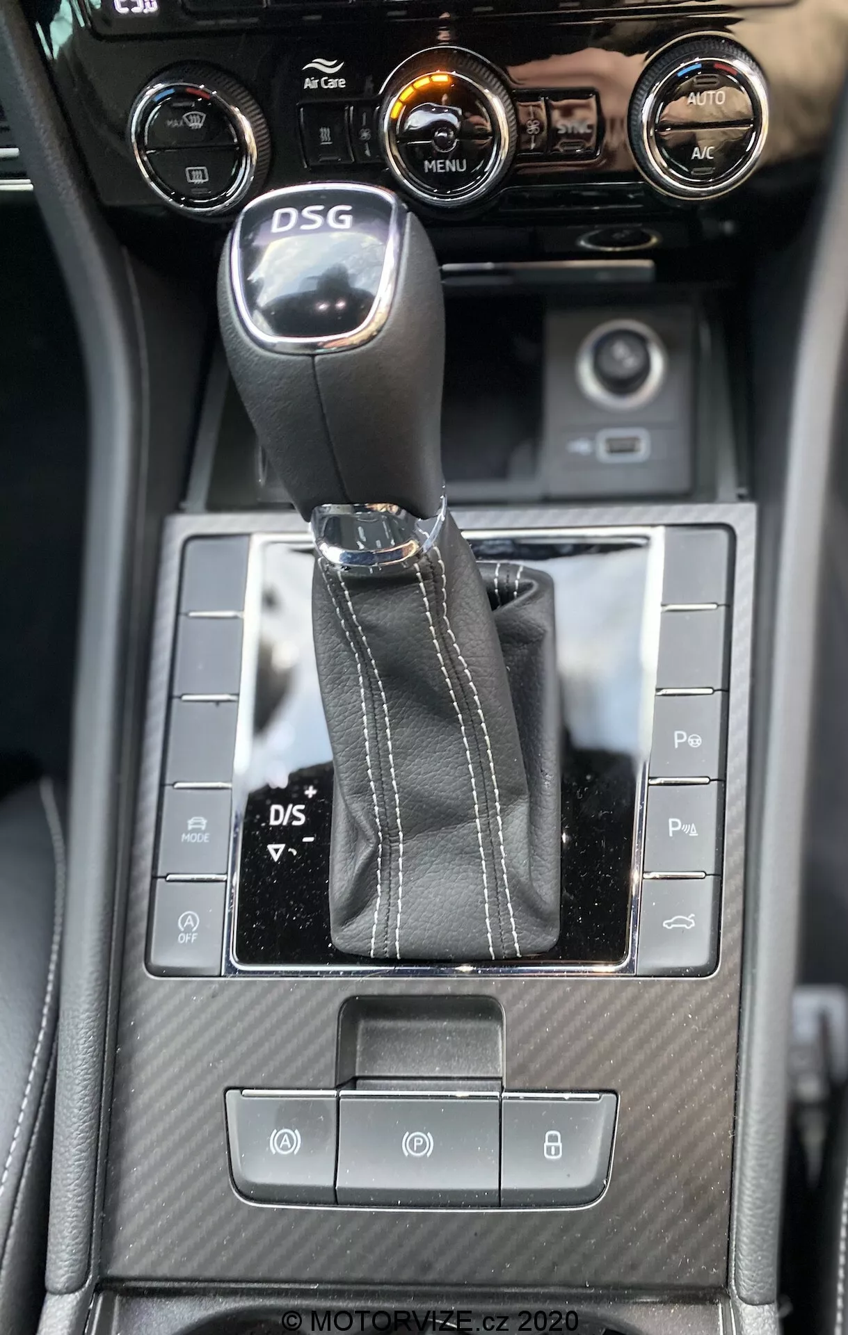 Widok wnętrza konsoli środkowej Skoda Superb 2019 z kąta skierowanego w dół, uwydatniający dźwignię zmiany biegów DSG, symetryczne przyciski sterujące, teksturowane detale przypominające włókno węglowe oraz regulatory klimatyzacji z centralnym ekranem dotykowym wyświetlającym informacje o pojeździe.
