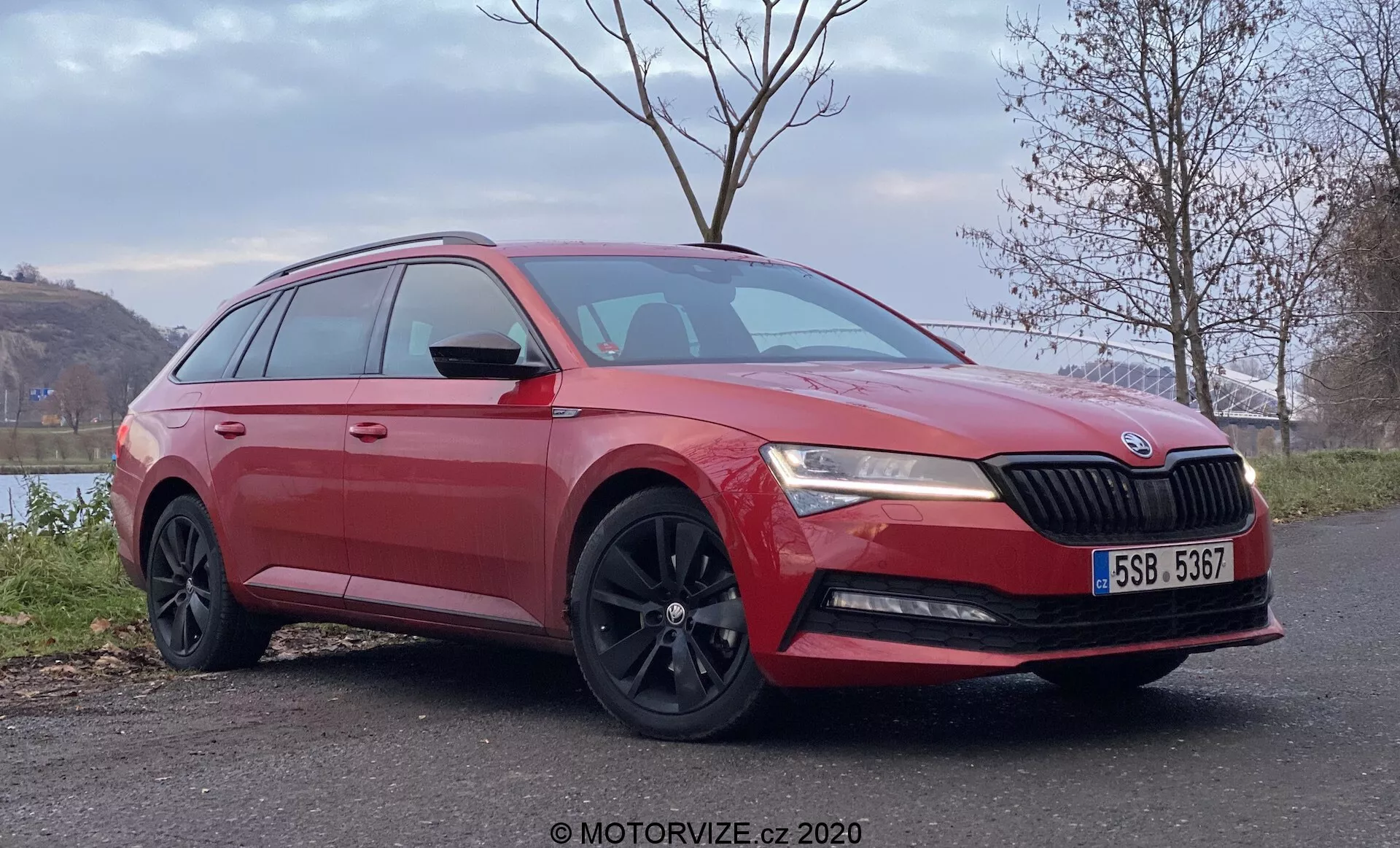 Obraz czerwonego kombi Škoda Superb widzianego z przodu pod kątem trzy czwarte, ukazującego charakterystyczny design grilla z emblematem Škoda, reflektory LED, rzeźbioną maskę, światła przeciwmgielne, wieloramienne czarne felgi aluminiowe, przyciemniane szyby i lusterka boczne z wbudowanymi kierunkowskazami, umieszczone w spokojnym wiejskim otoczeniu z roślinnością i wodą w tle.