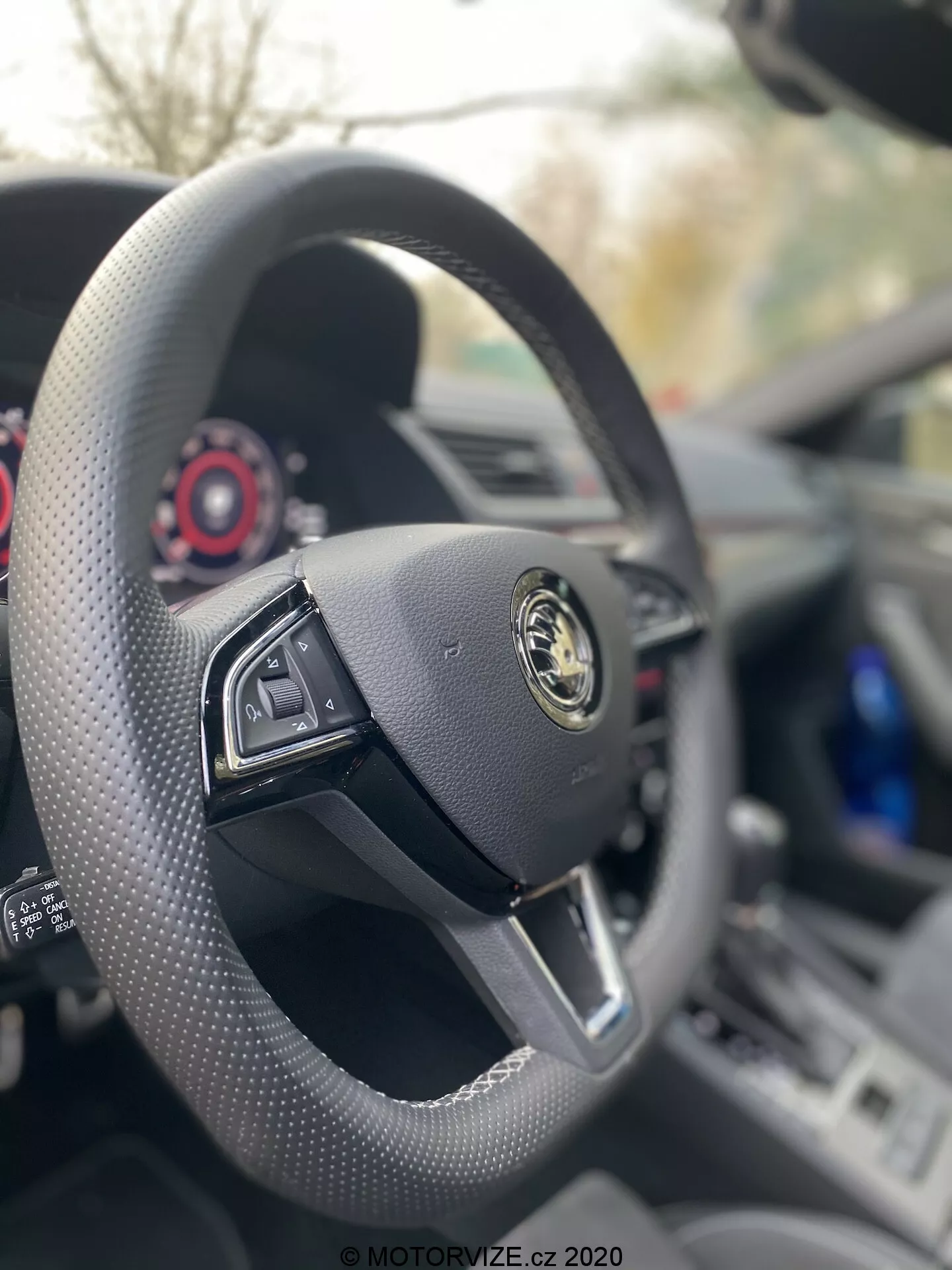 Wnętrze Škody Superb Limousine 2019 z perspektywy fotela kierowcy, skoncentrowane na kierownicy pokrytej czarną skórą, o płaskim dole z funkcjami wielofunkcyjnymi. Logo producenta, ze złotym emblematem na czarnym tle, jest wyraźnie widoczne na środku. Za kierownicą widoczna jest deska rozdzielcza i zestaw wskaźników, podkreślające minimalistyczny, luksusowy design auta z czerwonymi detalami na współczesnych tarczach.