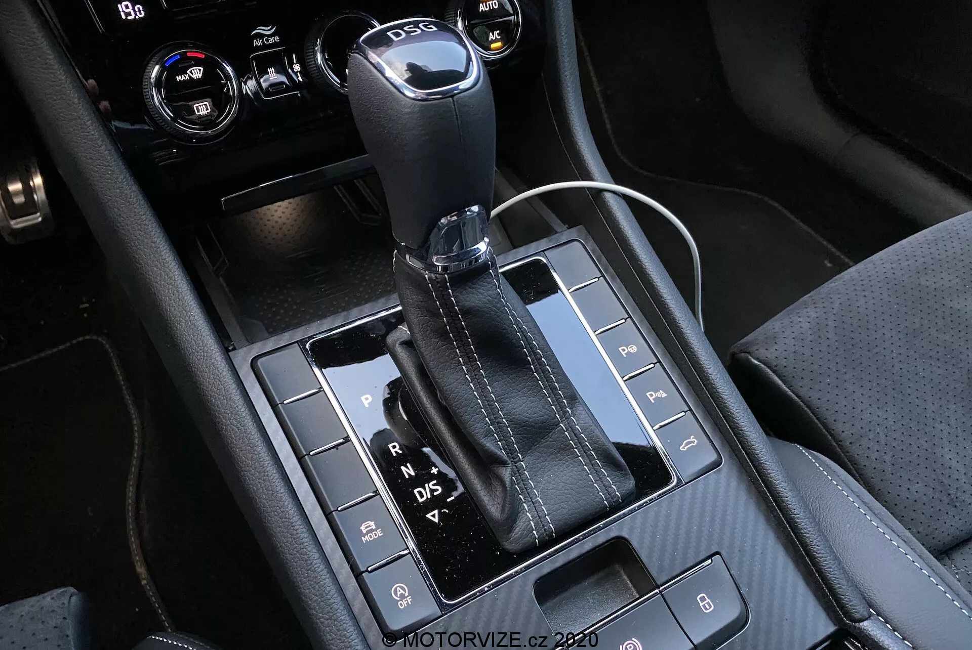 Zbliżenie na automatyczną dźwignię zmiany biegów i okolicę konsoli środkowej samochodu Skoda Superb 2019, z dźwignią zmiany biegów pokrytą skórą z metalową górą i widocznymi literami 'P, R, N, D/S' oznaczającymi różne biegi. Błyszczący czarny panel zawiera różne przyciski odpowiadające za funkcje takie jak czujniki parkowania, dezaktywacja systemu Auto Start/Stop i kontrola trakcji. Konsola łączy materiały o szczegółowym przeszyciu, w tym gniazdo zasilania 12V oraz prawdopodobnie pokrętła do sterowania klimatyzacją lub systemami multimedialnymi w przyciętym prawym górnym rogu.